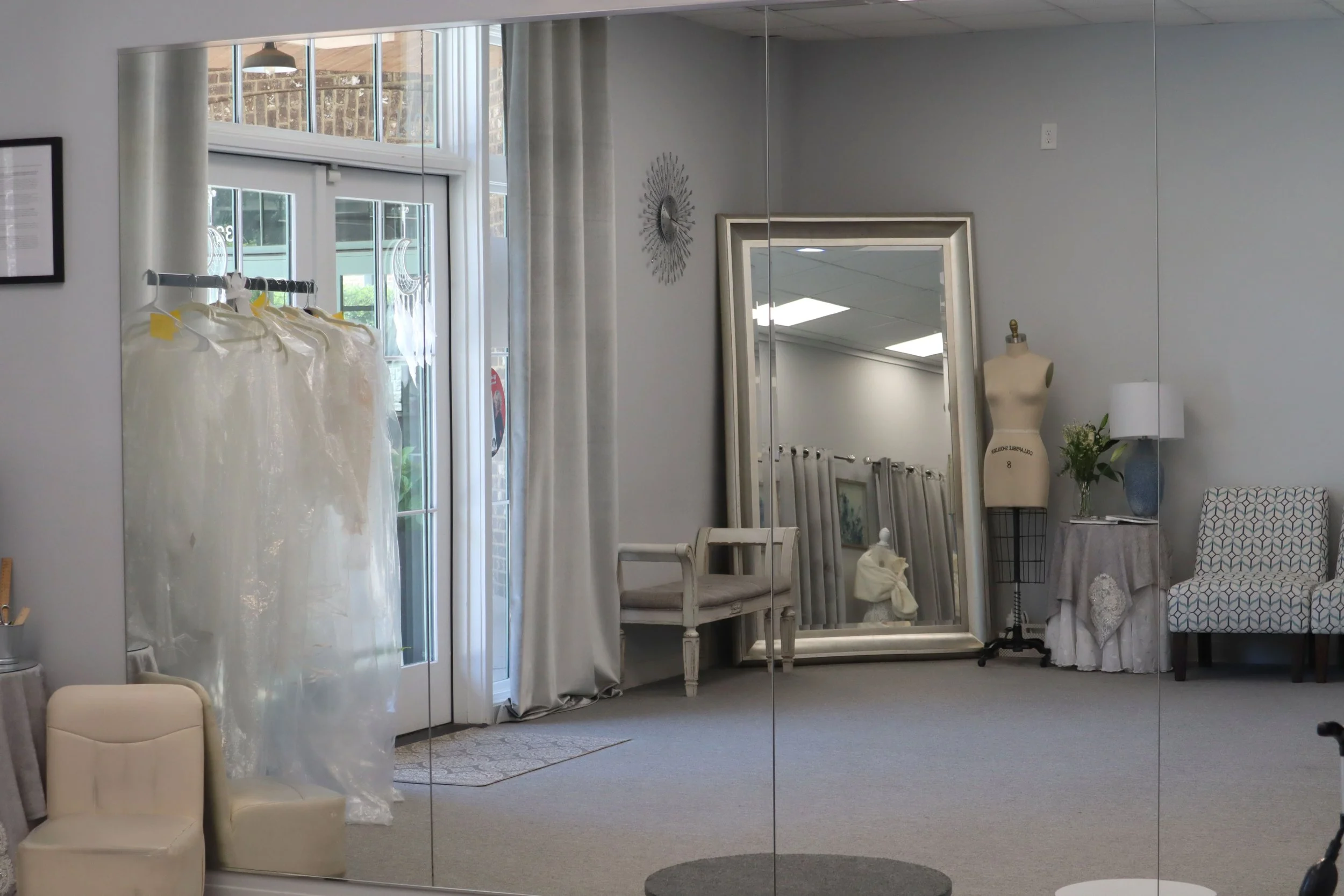 Interior view of a room with a large mirror reflecting a mannequin, a table with a lace tablecloth, and a potted plant. There are chairs, curtains, and a window with a view outside.