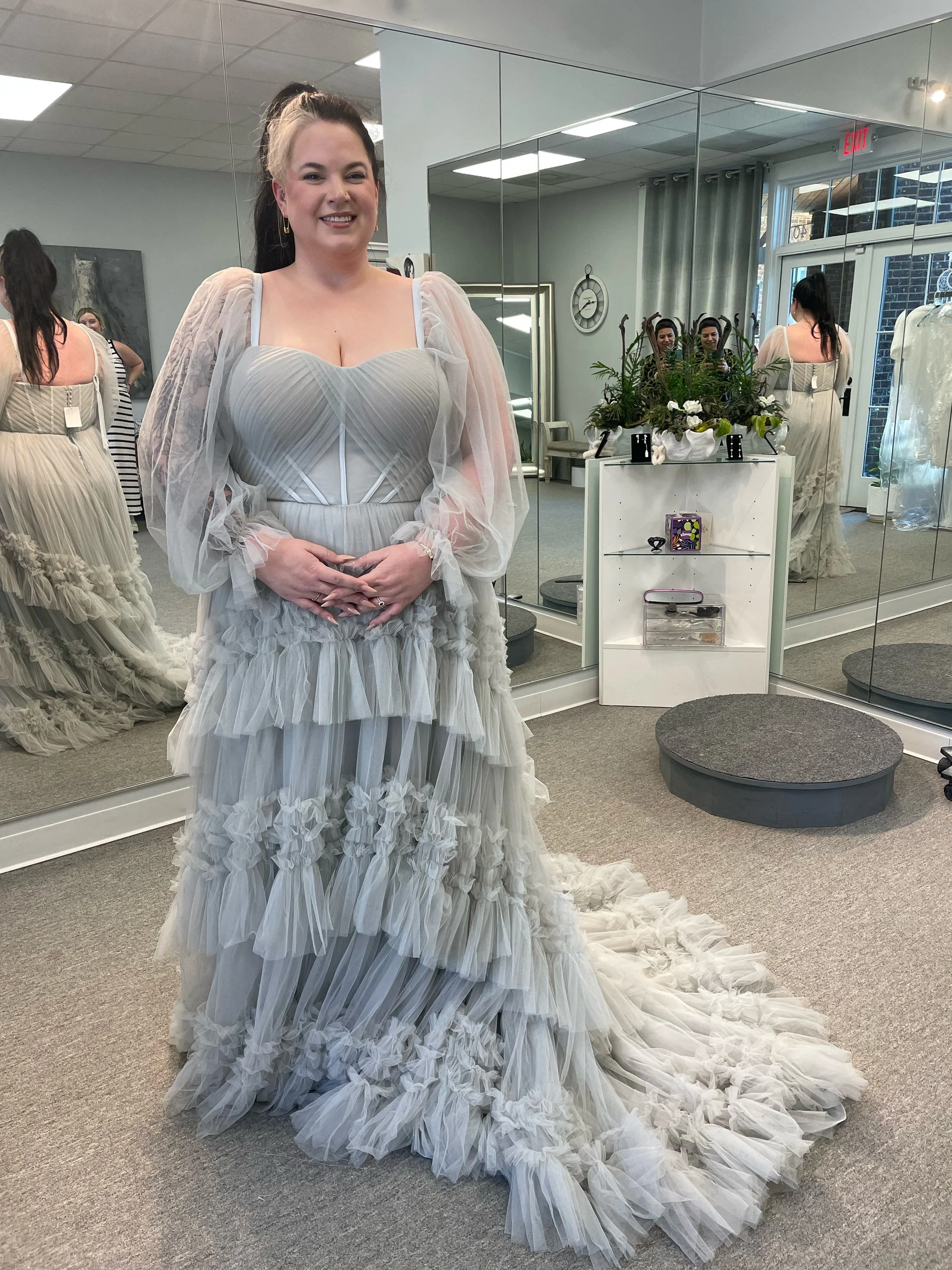 A woman wearing a white wedding dress with layers of ruffled tulle, standing in front of a large mirror inside a bridal boutique. The dress has puffed sheer sleeves and a fitted bodice. The train of the dress flows onto the floor, and the woman is smiling with her hands clasped in front of her. There are multiple people behind her, trying on dresses, and the boutique has a modern decor with a clock on the wall, plants, and a small white shelf.