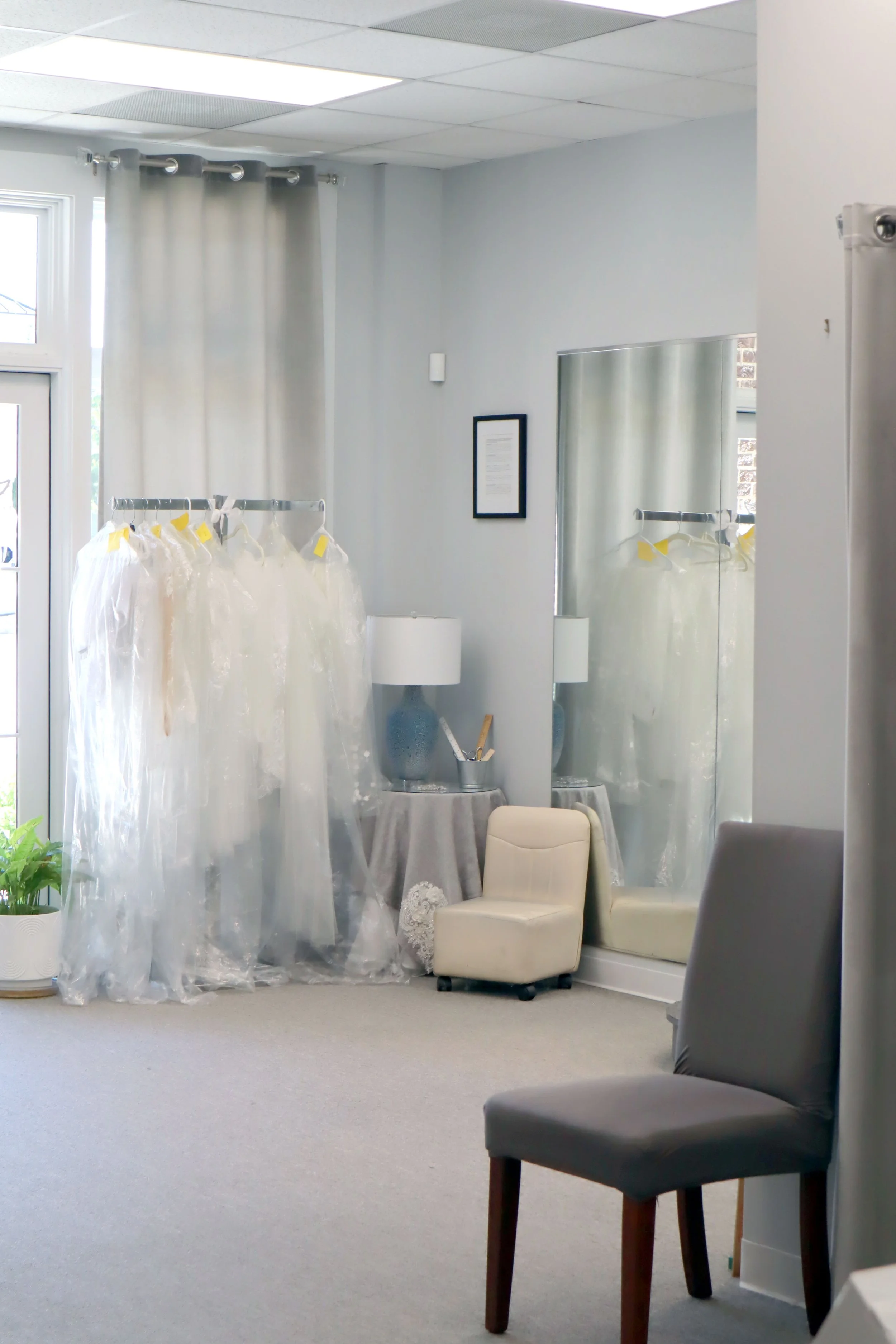 A room set up for a wedding or event, with bridal gowns on a clothing rack, a mirror, chairs, a small round table with a lace cloth, and decorative lamps. Light gray walls, ceiling tiles, and windows with curtains are visible.