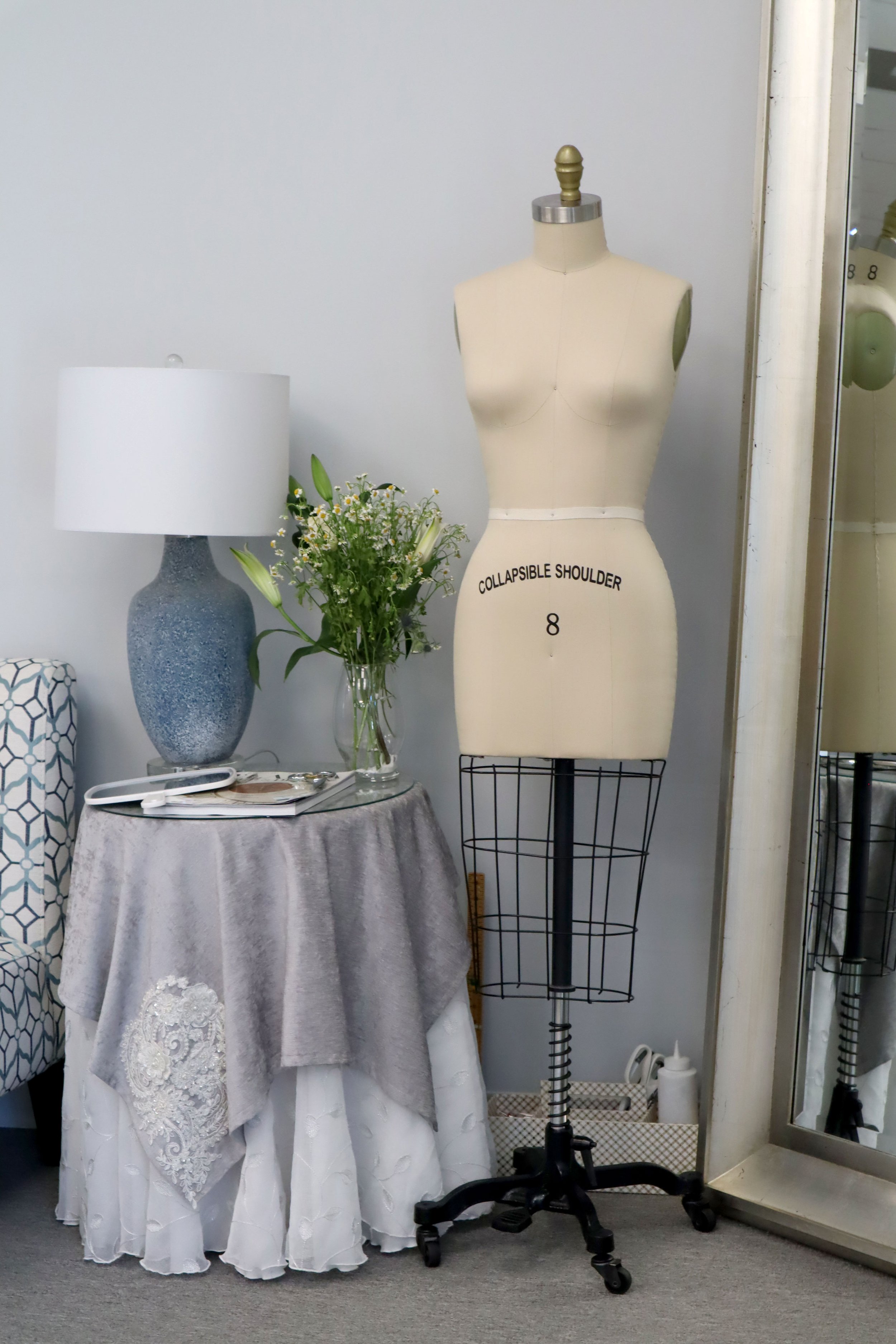 A dressmaker's mannequin marked 'COLLAPSIBLE SHOULDER 8' next to a table with a lace tablecloth, a vase of flowers, a lamp, and a sewing machine; a mirror on the right and a rolling cart with paper towels underneath.