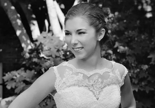 Black and white photo of a young woman in a wedding dress, smiling outdoors with trees and foliage in the background.