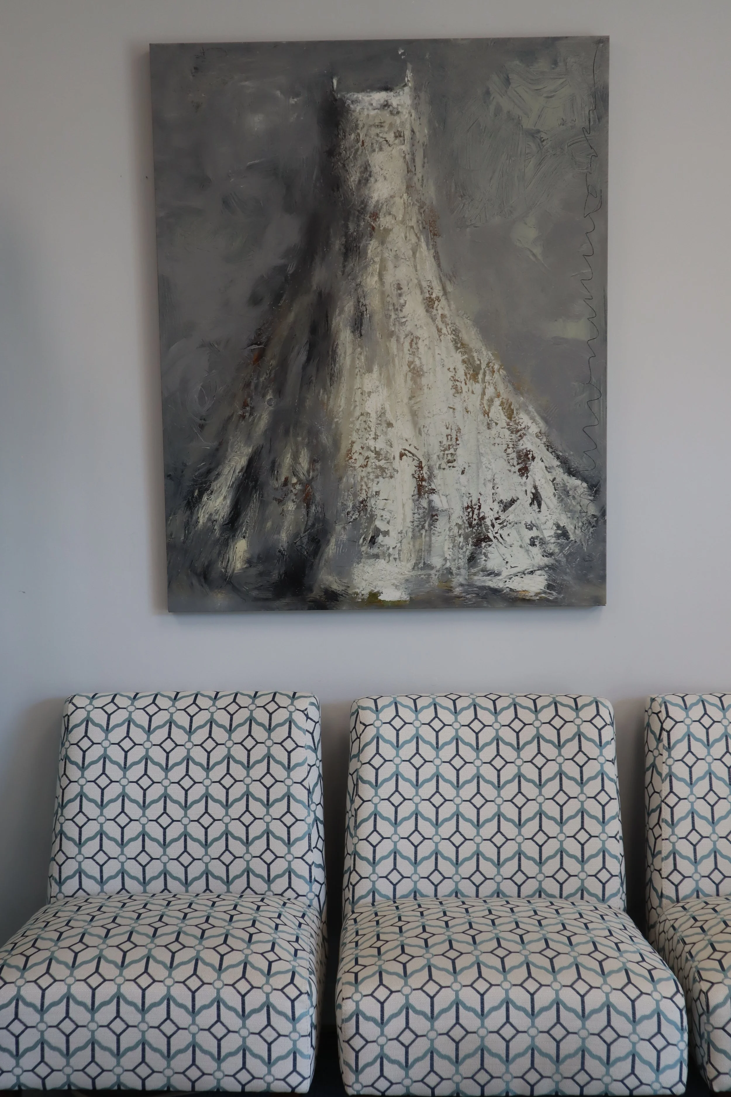 Three patterned chairs with a geometric design placed in front of a plain wall. Above the chairs hangs a large abstract painting with shades of gray, black, white, and beige.
