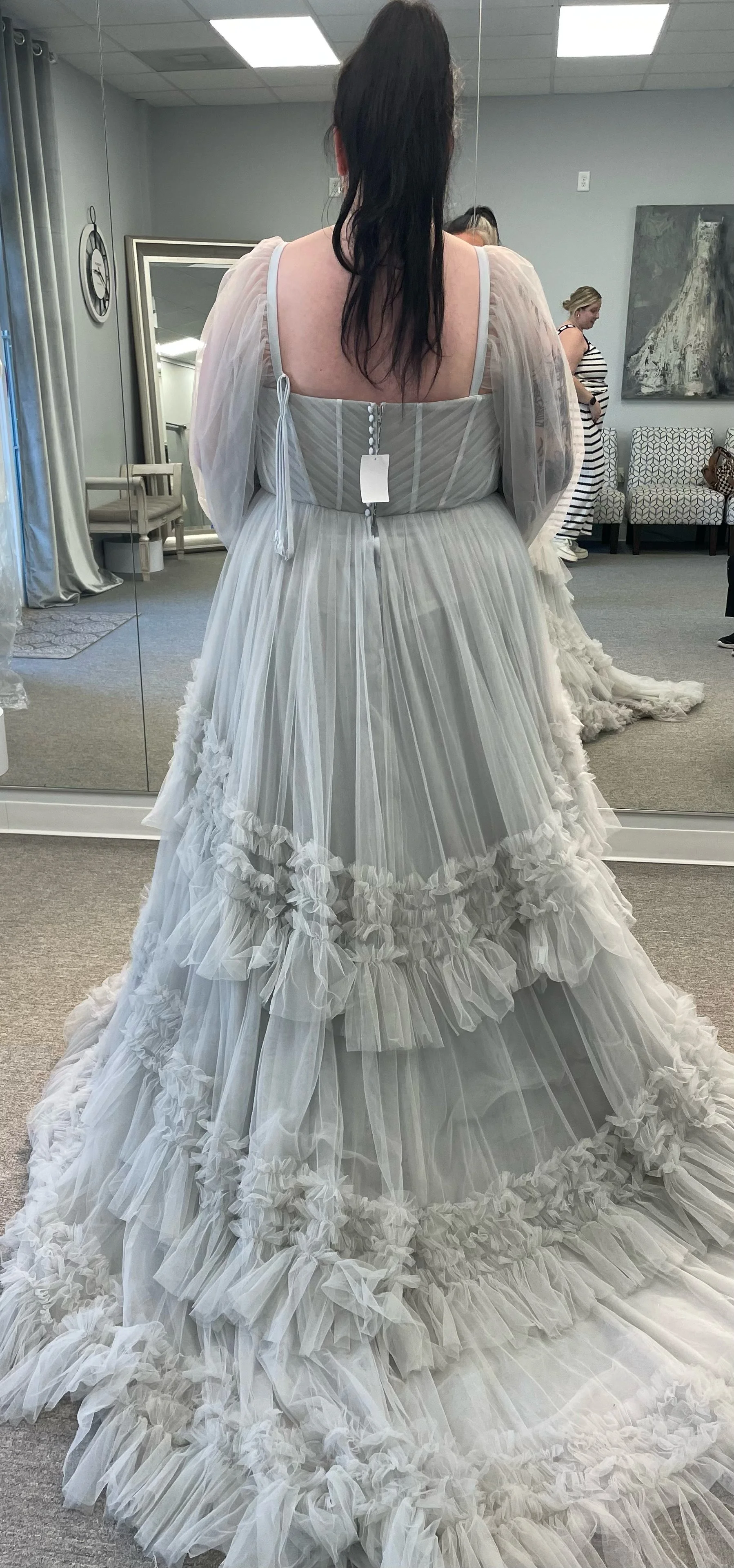 A woman trying on a white wedding dress with ruffles and sheer puffed sleeves, seen from behind in front of a mirror in a bridal boutique.