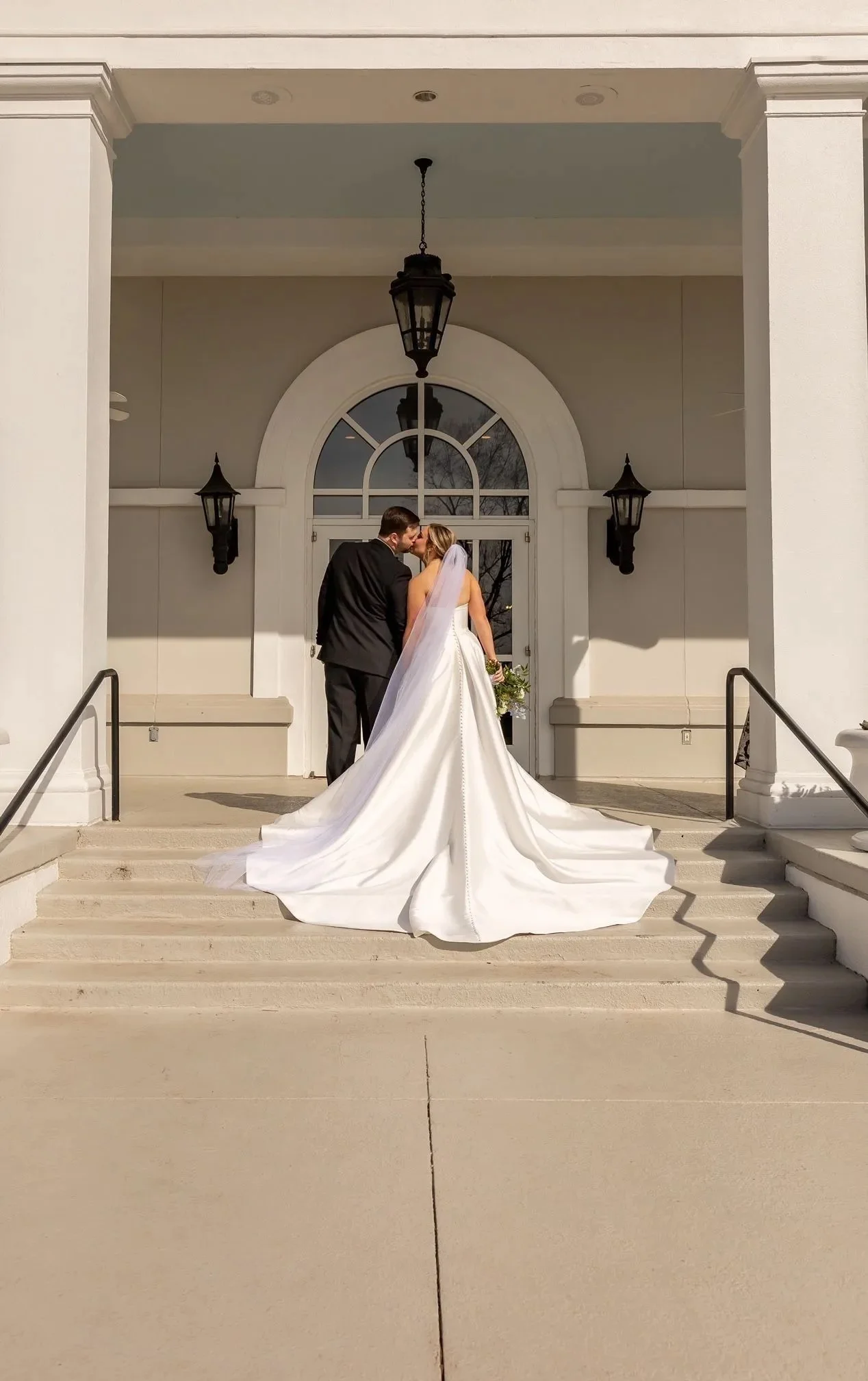 A bride and groom share a kiss on the steps in front of a large white building with an arched window. The bride is in a white wedding gown with a long train and veil, holding a bouquet. The groom is in a black suit. Two black lantern-style light fixtures are mounted on either side of the arched window.
