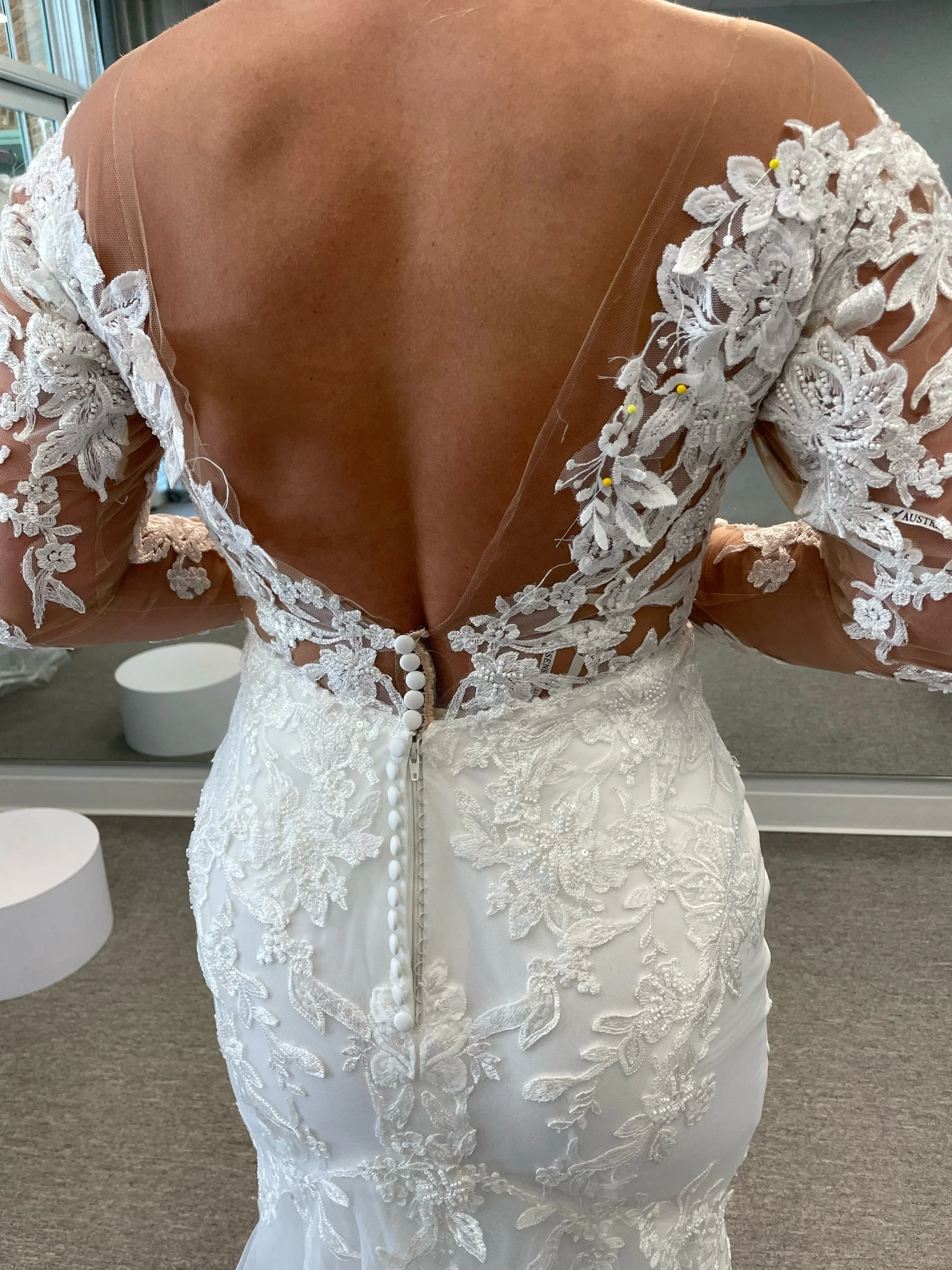 Back view of a woman in a white wedding dress with lace floral details, buttons down the back, and sheer lace sleeves with floral embroidery.