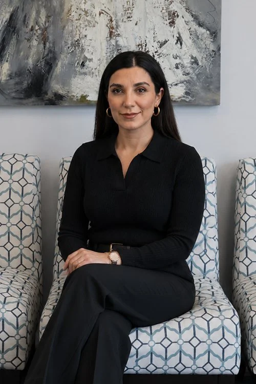 A woman with long dark hair, wearing a black long-sleeve top and black pants, seated on a patterned armchair in front of an abstract painting.