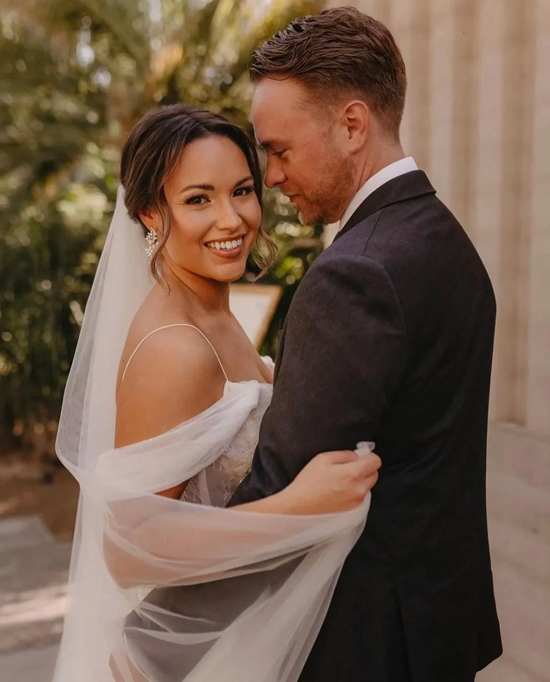 A bride and groom are embracing outdoors, sharing a joyful moment on their wedding day. The bride is smiling at the camera while the groom has his eyes closed, leaning into her. The bride is wearing a white wedding dress and veil, with earrings, and has her arm around the groom. The groom is dressed in a dark suit and white shirt.