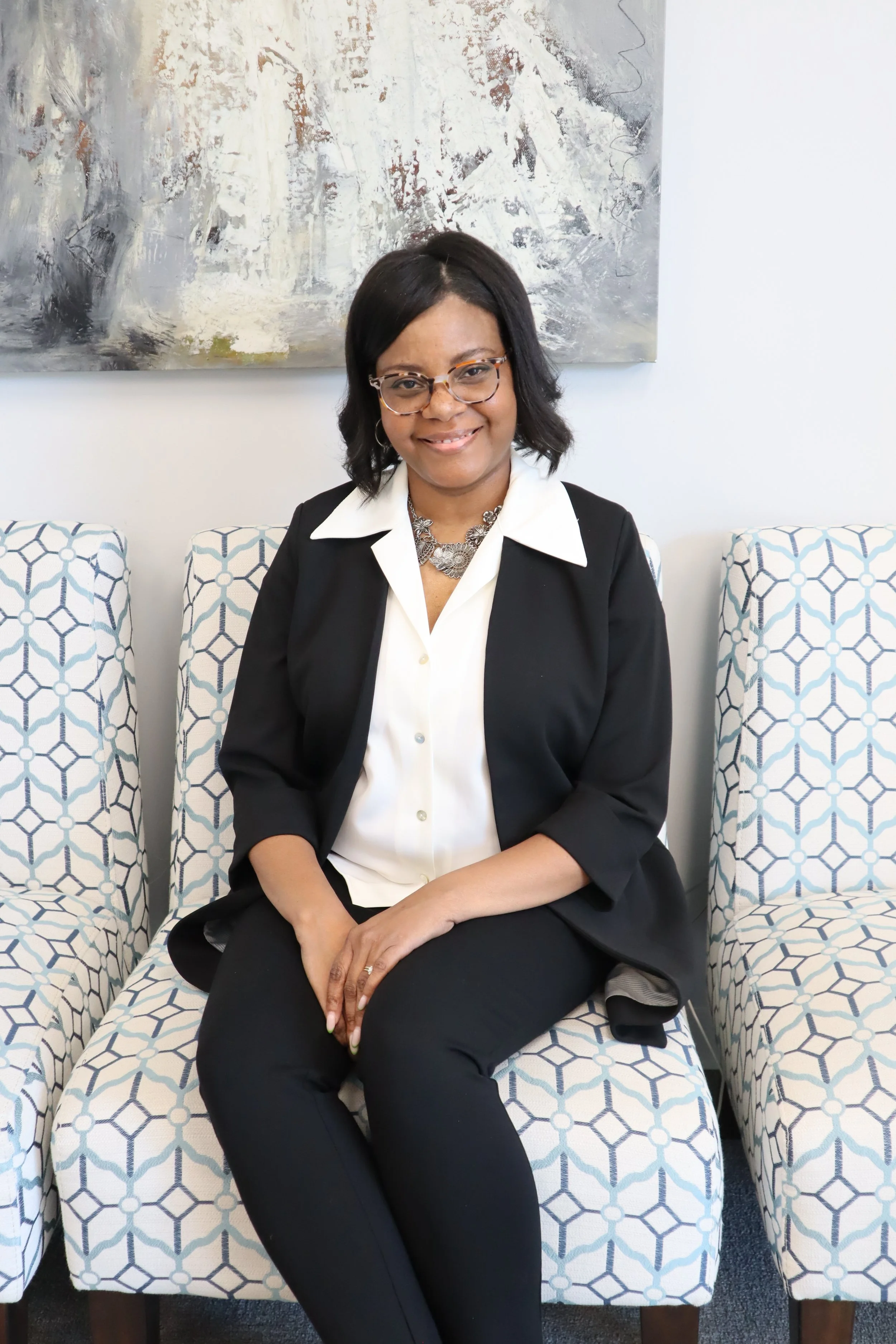 A woman with glasses, wearing a black blazer and white shirt, sitting on a patterned chair in front of an abstract painting on a white wall.