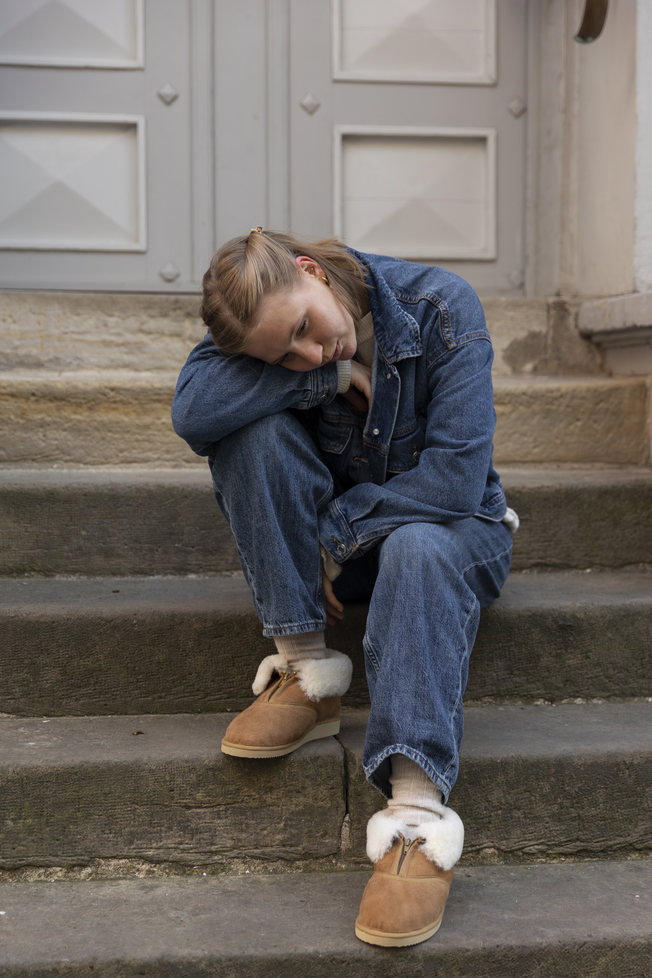 A young woman with long blonde hair sitting on outdoor stairs, resting her head on her knee, wearing a denim jacket, blue jeans, tan moccasin boots with white fur trim, and cream-colored socks.
