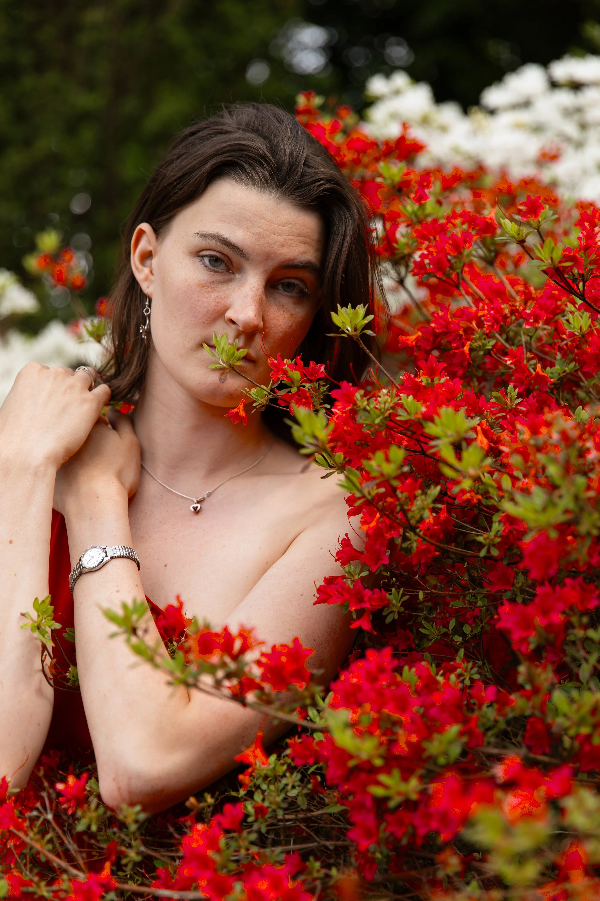 A woman with short dark hair and blue eyes, wearing jewelry including a necklace with a heart, earrings, and a watch, posing amidst vibrant red azalea flowers and greenery outdoors.