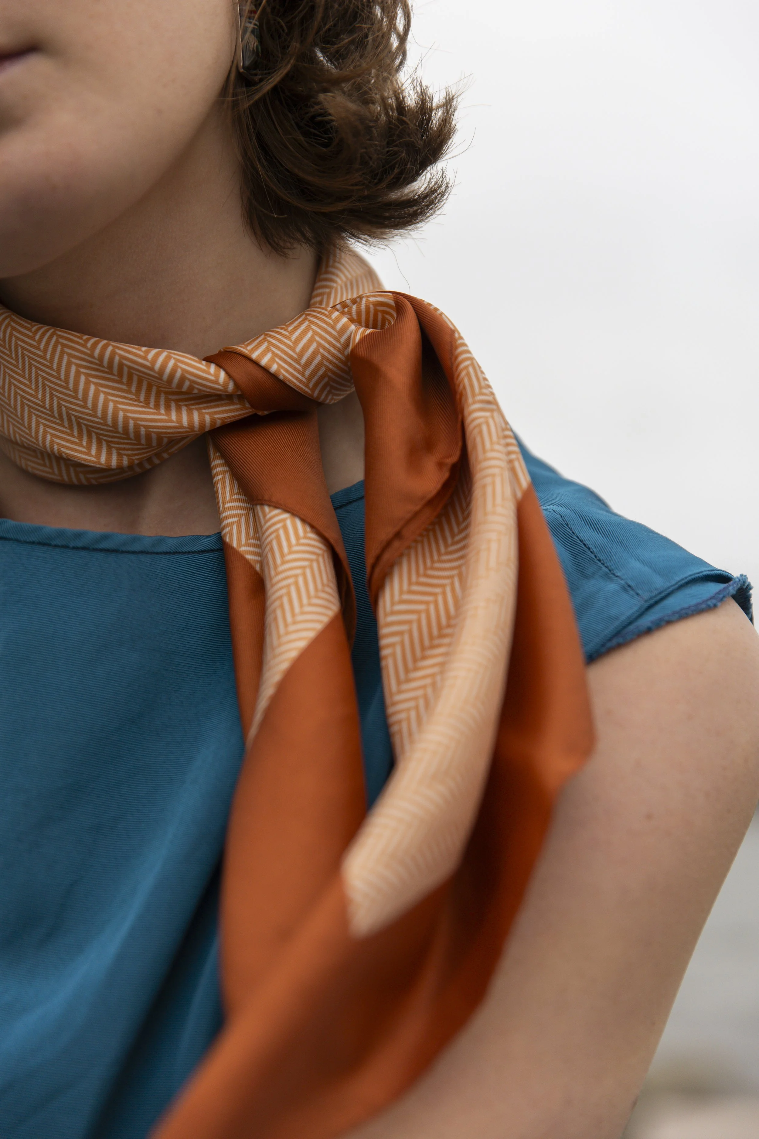 A close-up photo of a person wearing an orange patterned scarf, a blue shirt, and a face mask hanging from their ear.
