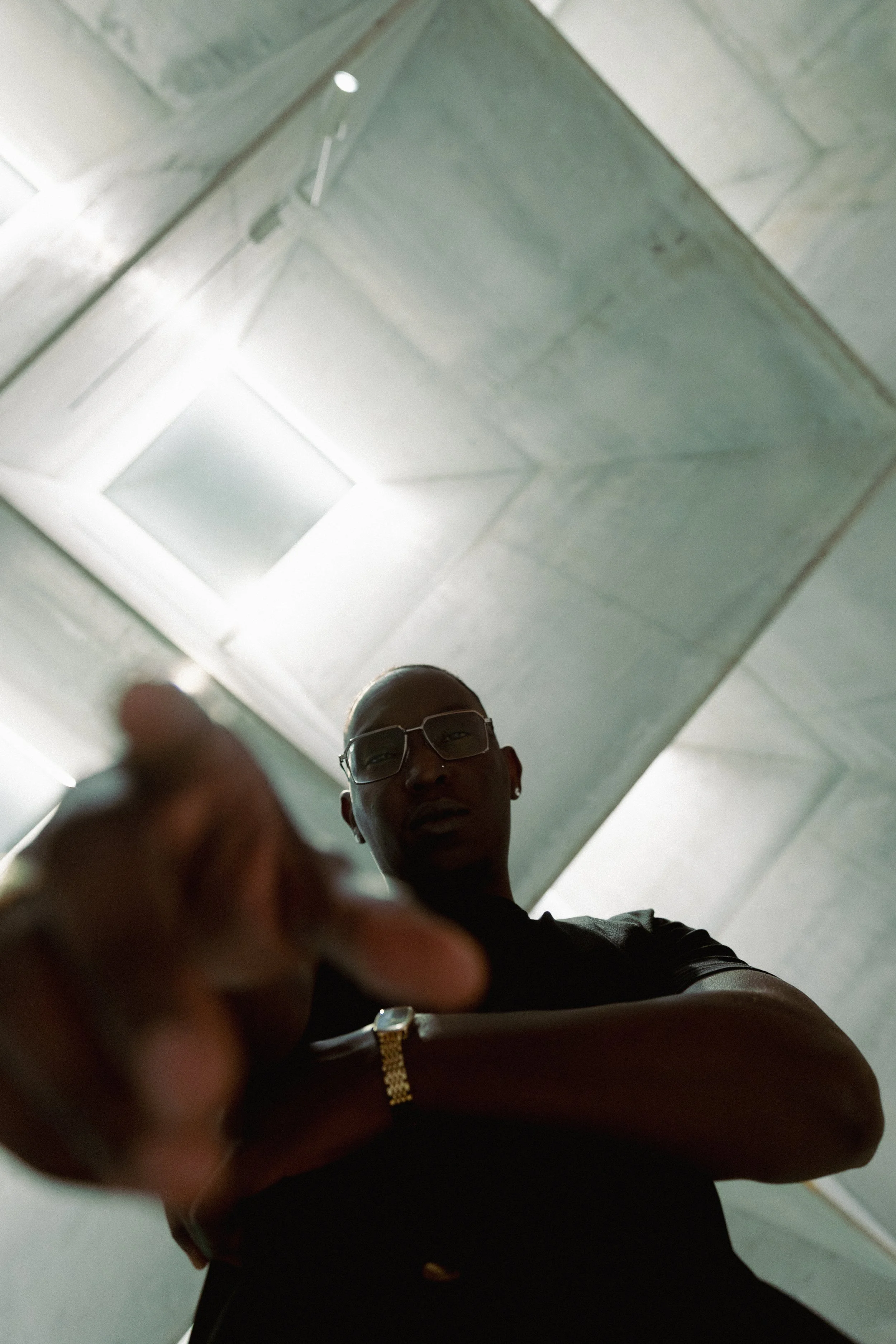 A person with glasses reaching towards the camera in a low-angle shot, with a concrete ceiling and lights visible above.