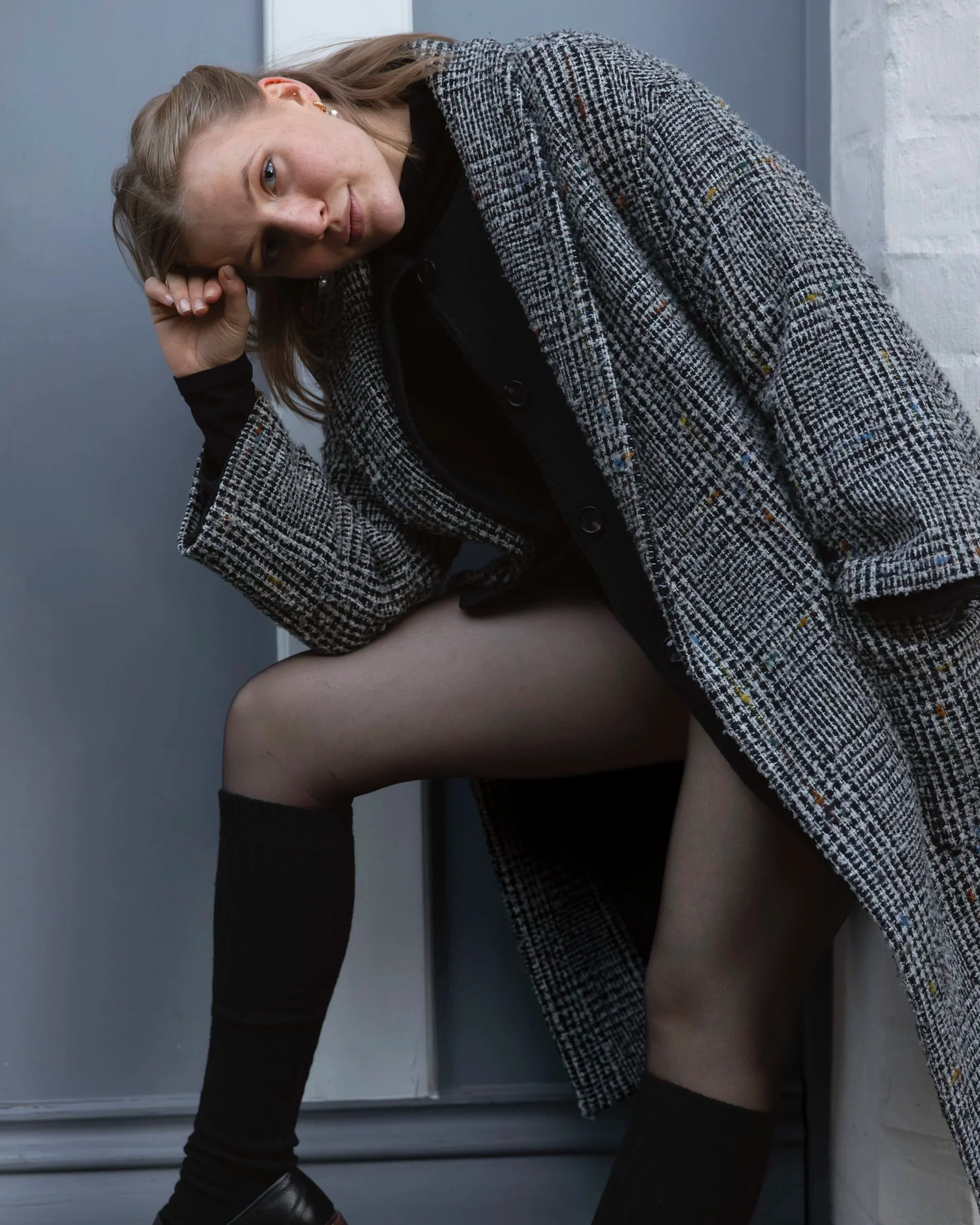 A woman with light skin and freckles, wearing a gray checkered coat, a black top, and beige tights, is sitting on a windowsill with her head resting on her hand, looking at the camera. The background features a light gray wall and window frame.