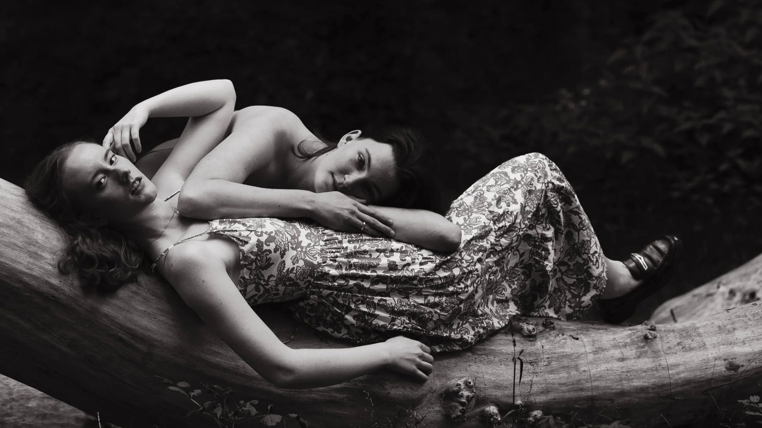 Two women lying on a log outdoors, one wearing a floral dress and the other with a dark top, both smiling and looking at the camera, black and white photo.