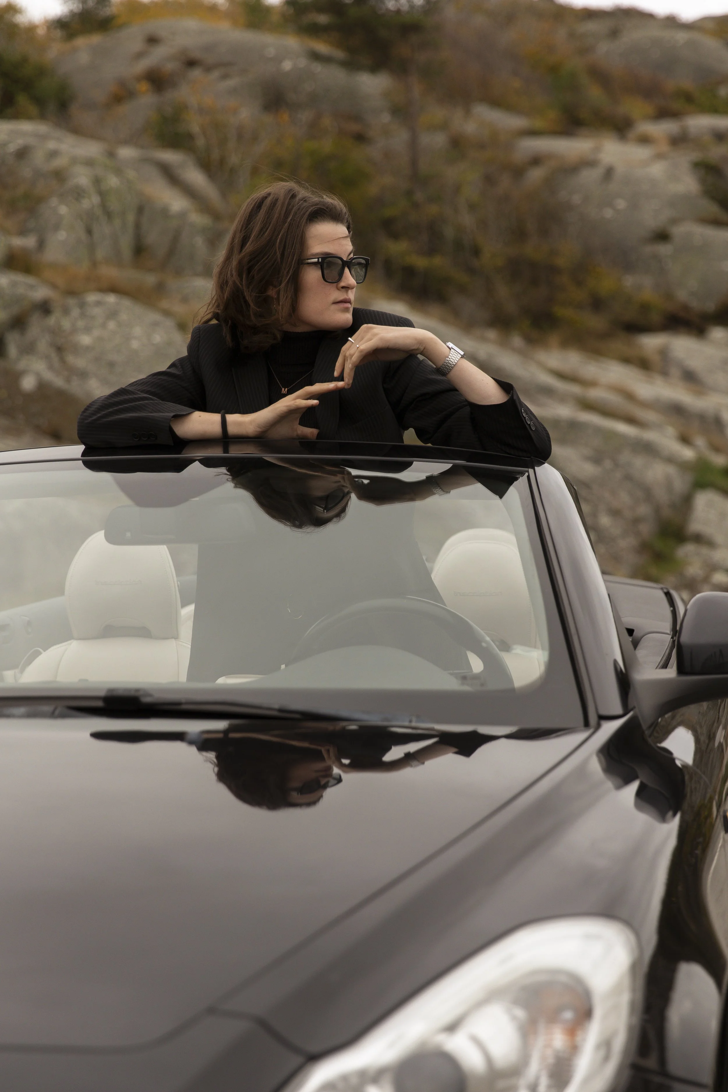 A woman with brown hair, wearing glasses, a black blazer, and jewelry, is leaning on the car roof with her arm resting while looking to the side. She is outdoors with rocky terrain and sparse trees in the background, captured through the reflection on a black convertible car's windshield.