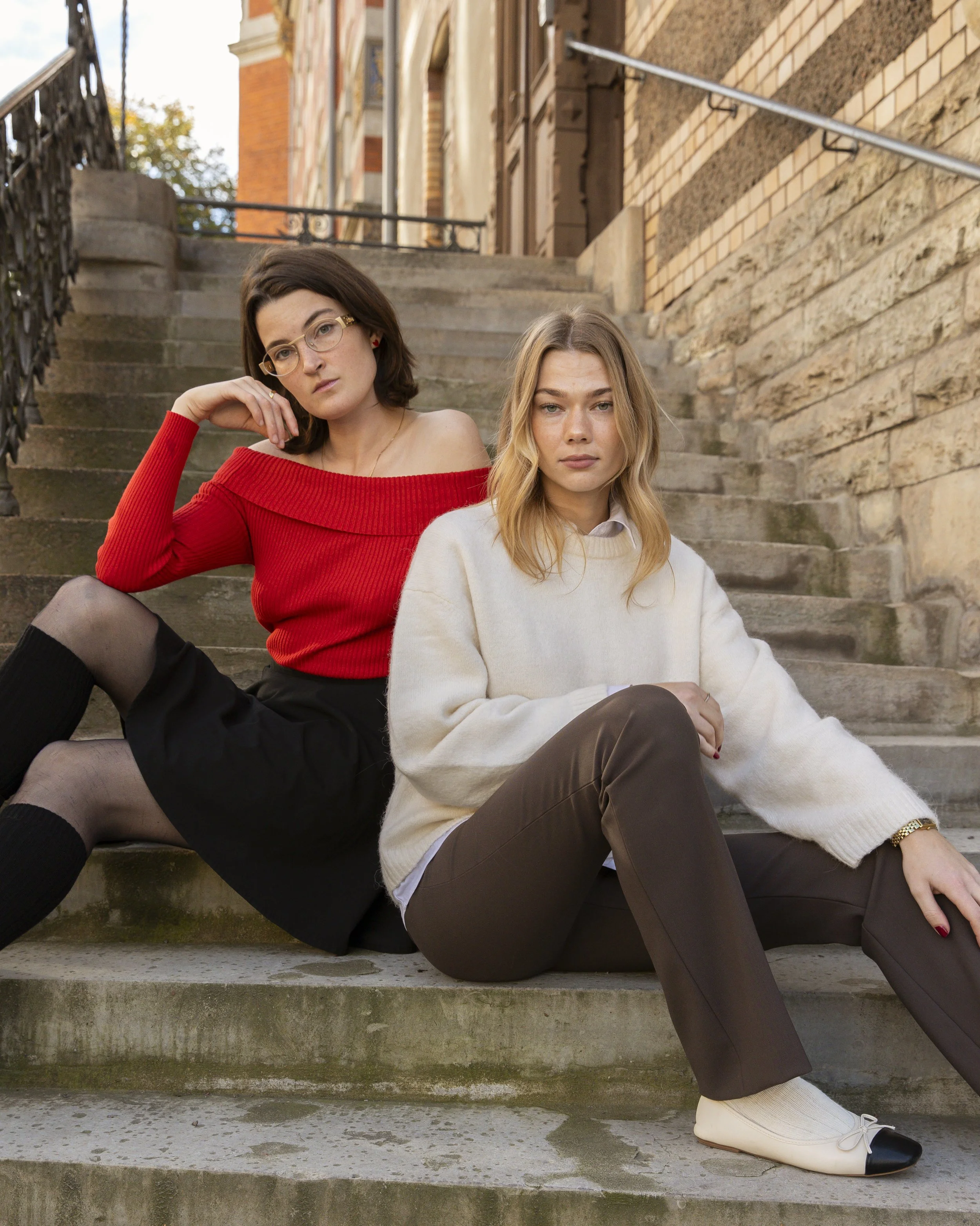 Two young women sitting on outdoor concrete stairs in front of a brick building. One woman has short brown hair, glasses, red off-shoulder sweater, and black skirt with sheer stockings. The other woman has long blonde hair, white sweater, and brown pants. They appear to be posing for a photo.
