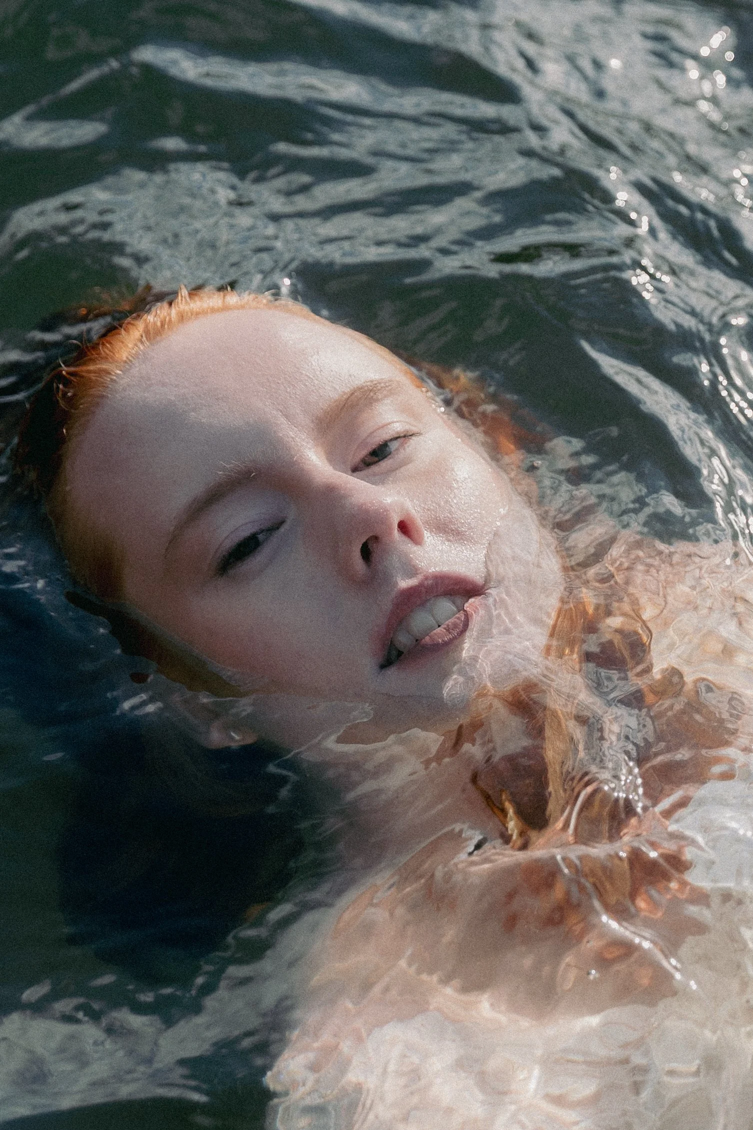 A person with red hair swimming in water, with only their face visible as they look upward.