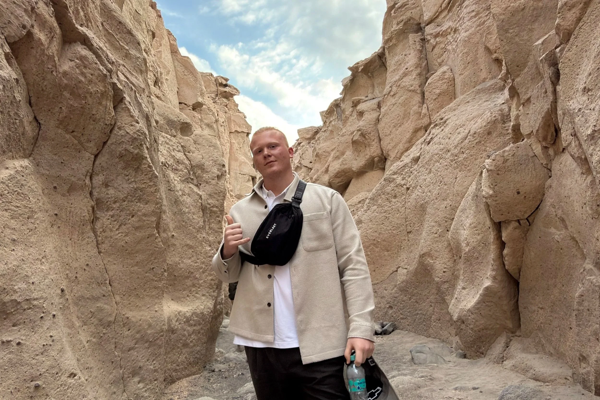 A young man with short blonde hair standing in a narrow canyon with large beige stone walls on either side, under a partly cloudy sky. He is wearing a light-colored jacket, a white shirt, and has a black crossbody bag. He is holding a water bottle in one hand and giving a thumbs-up with the other.