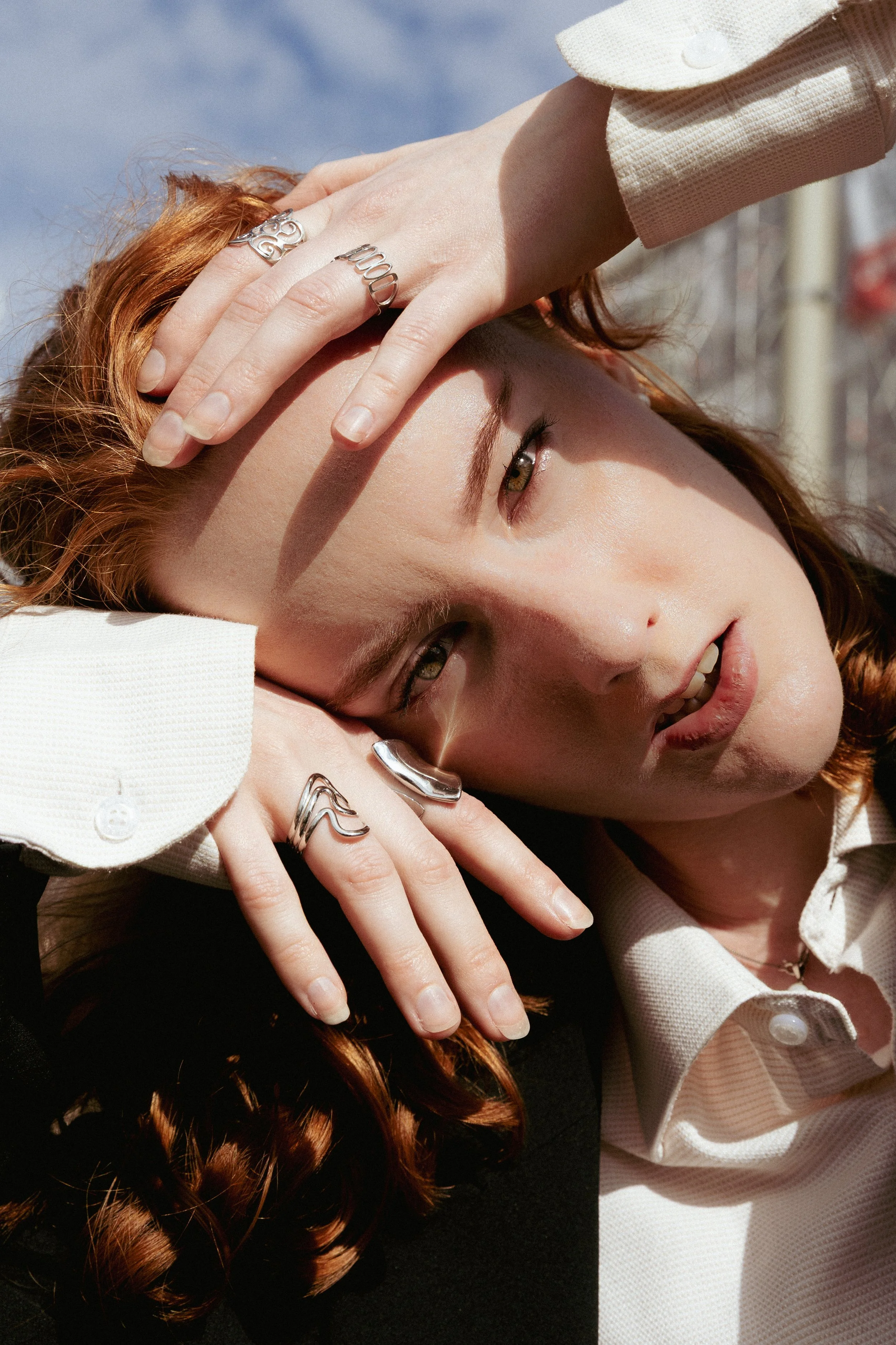 A woman with red hair lying on her side, resting her head on her hand, wearing multiple silver rings and a beige jacket with white buttons.