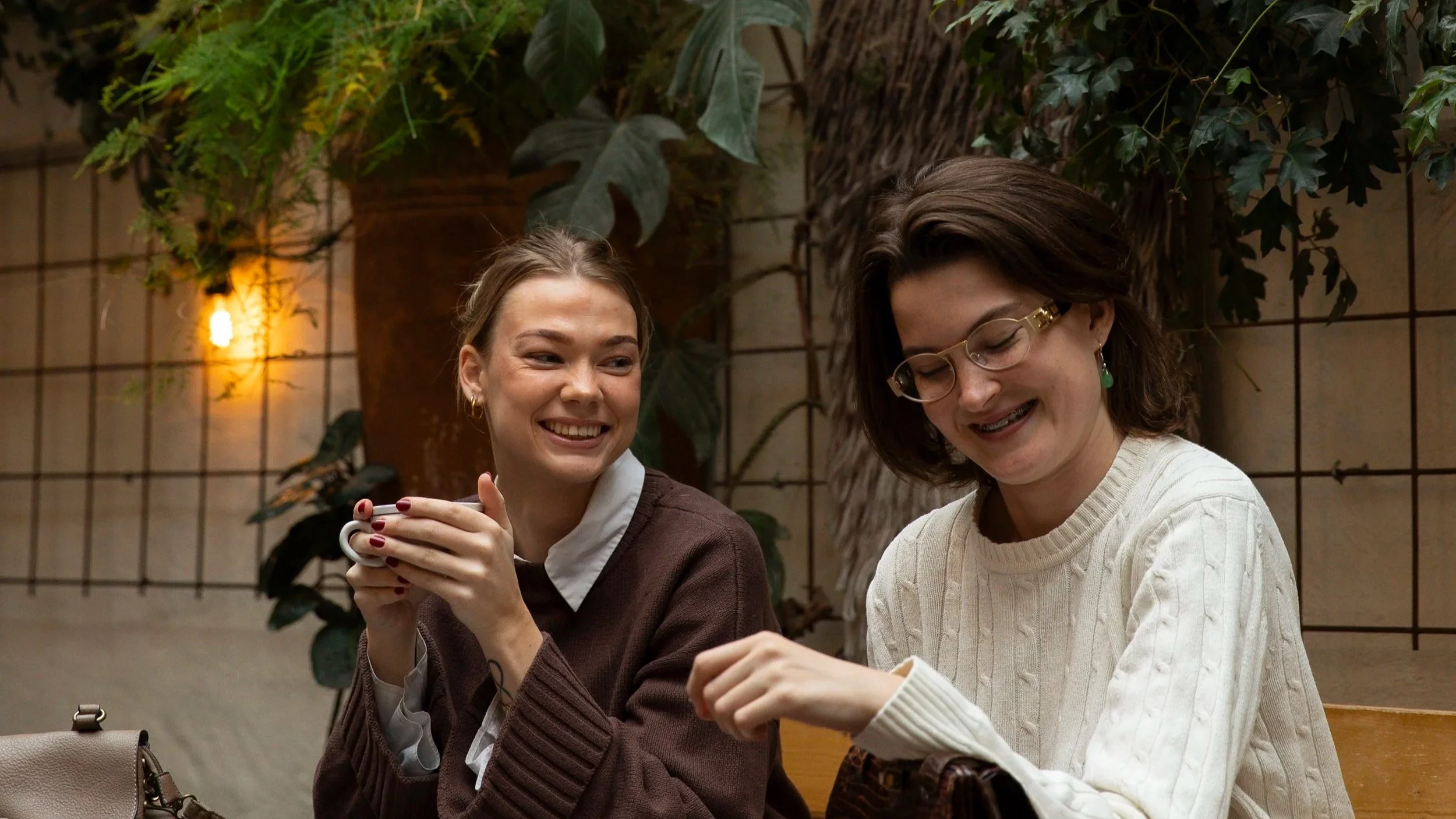 Two women sitting at a table in a cozy indoor space with plants, smiling and enjoying conversation. One woman is holding a mug.