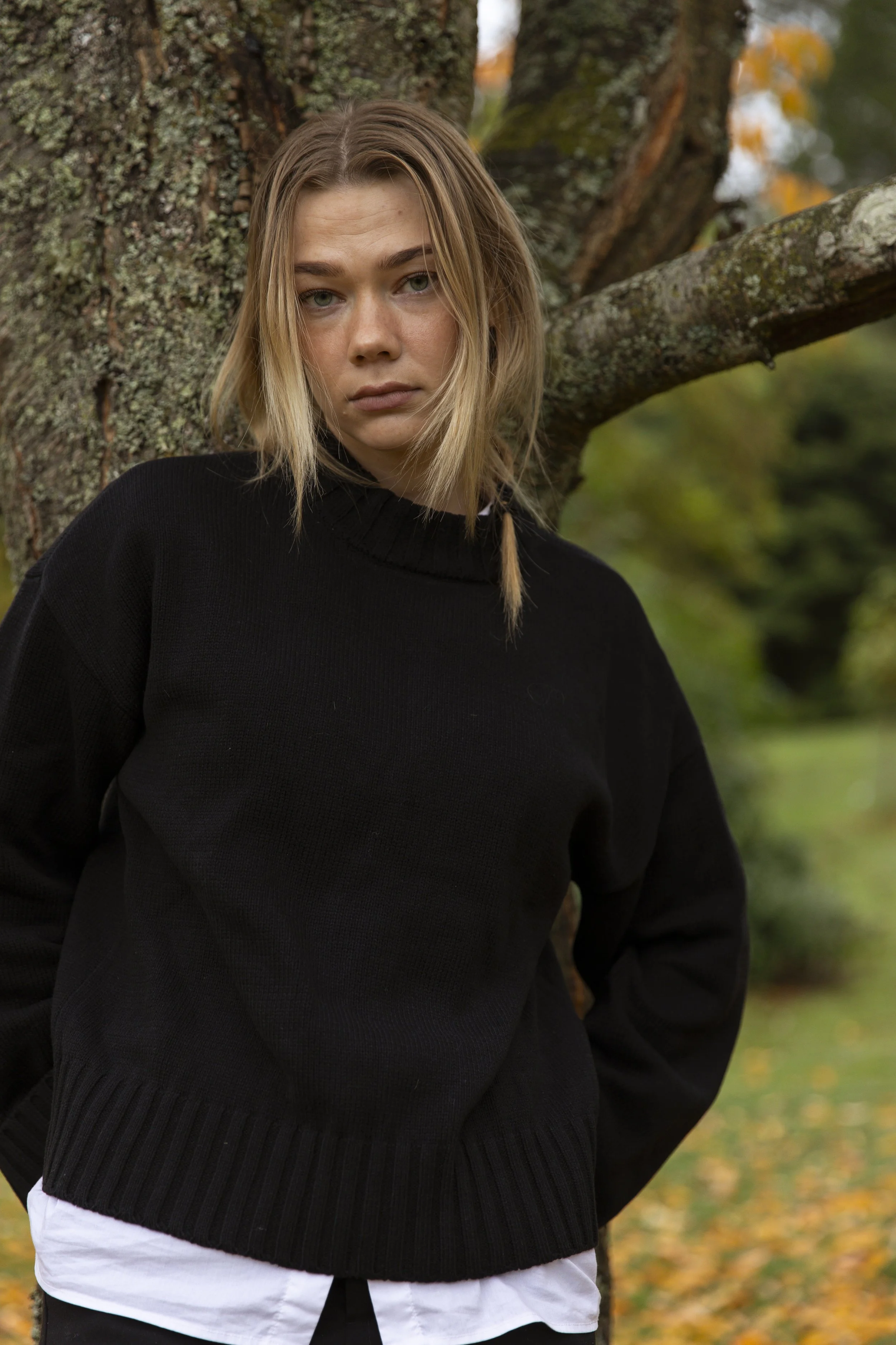 A young woman with blonde hair and blue eyes standing outdoors by a tree in a park, wearing a black sweater and a white shirt underneath, with autumn leaves on the ground.