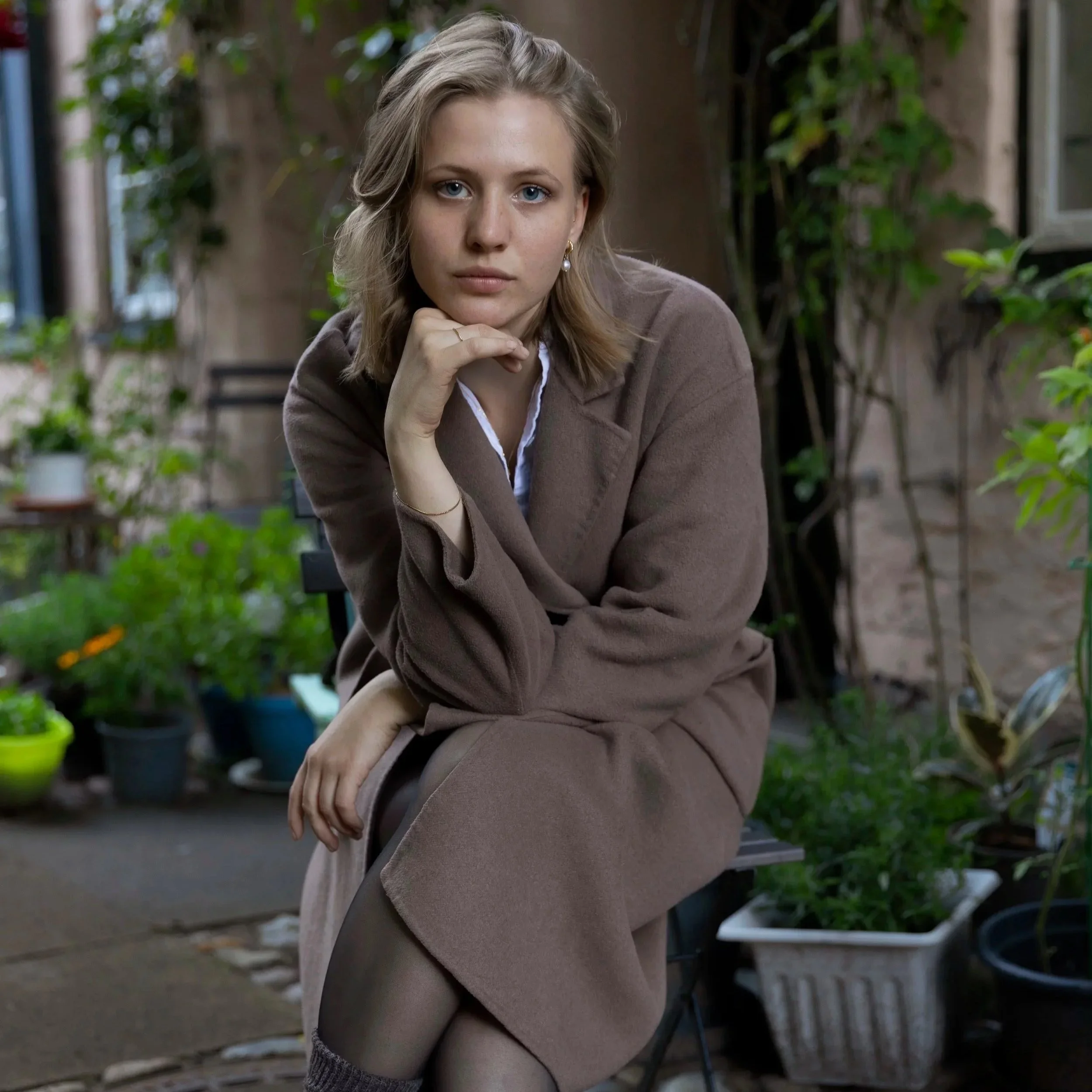 A young woman with blonde hair and blue eyes sitting outdoors among green plants, wearing a beige blazer and earrings, with her hand resting on her chin, looking pensively at the camera.