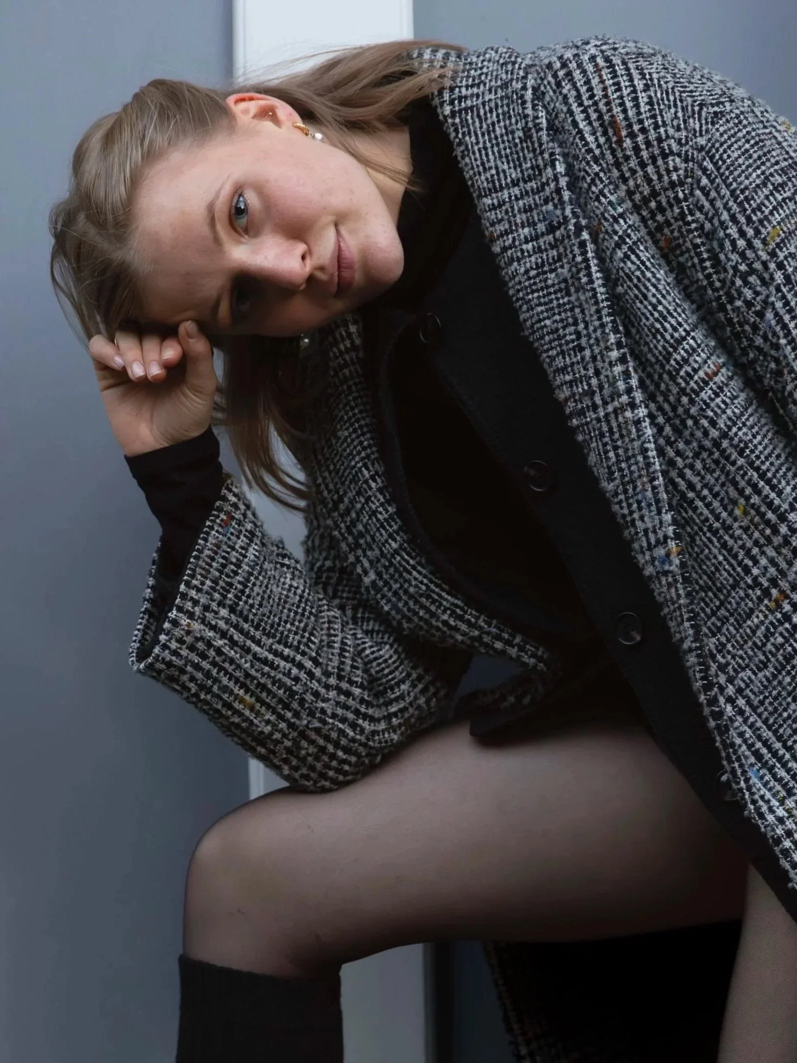 A woman with light-colored hair lies on her side on a bed, wearing a checkered jacket and looking into the camera. The background is a plain wall.