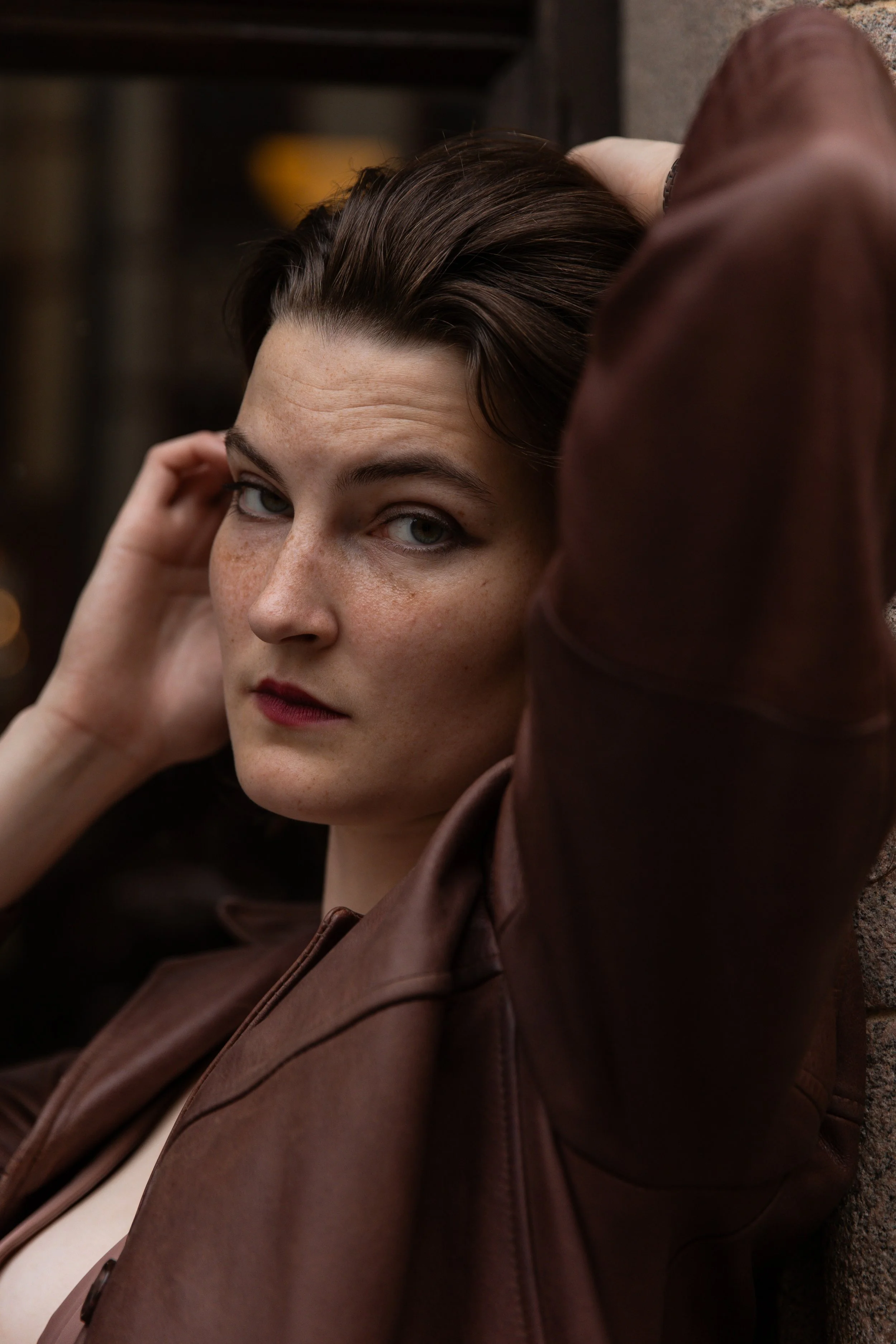 A woman with short brown hair and blue eyes resting her head against a brick wall, wearing a brown leather jacket.