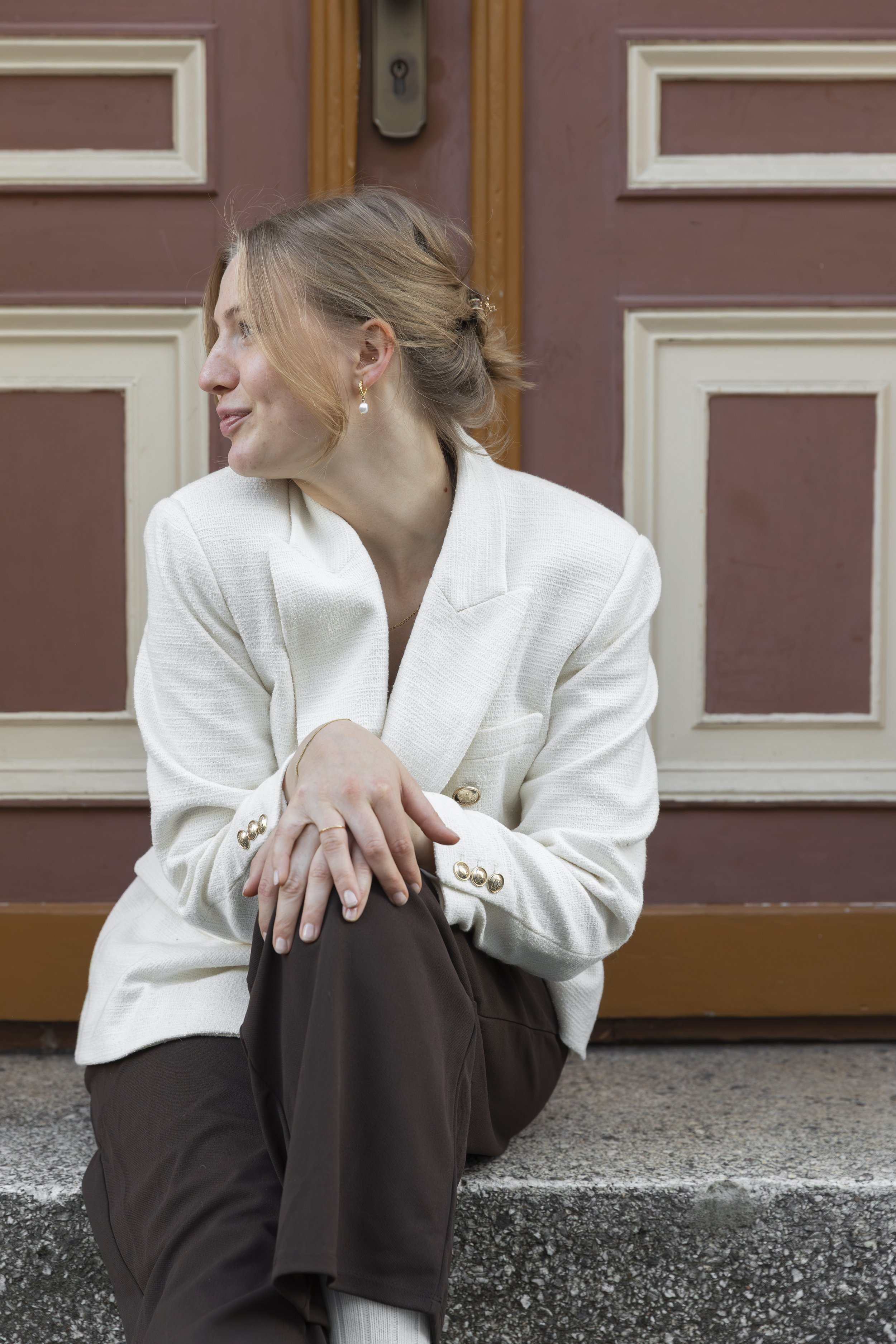 A woman with blonde hair in a loose bun wearing earrings and a white blazer, sitting on a cement step in front of a brown wooden door with decorative white trim.