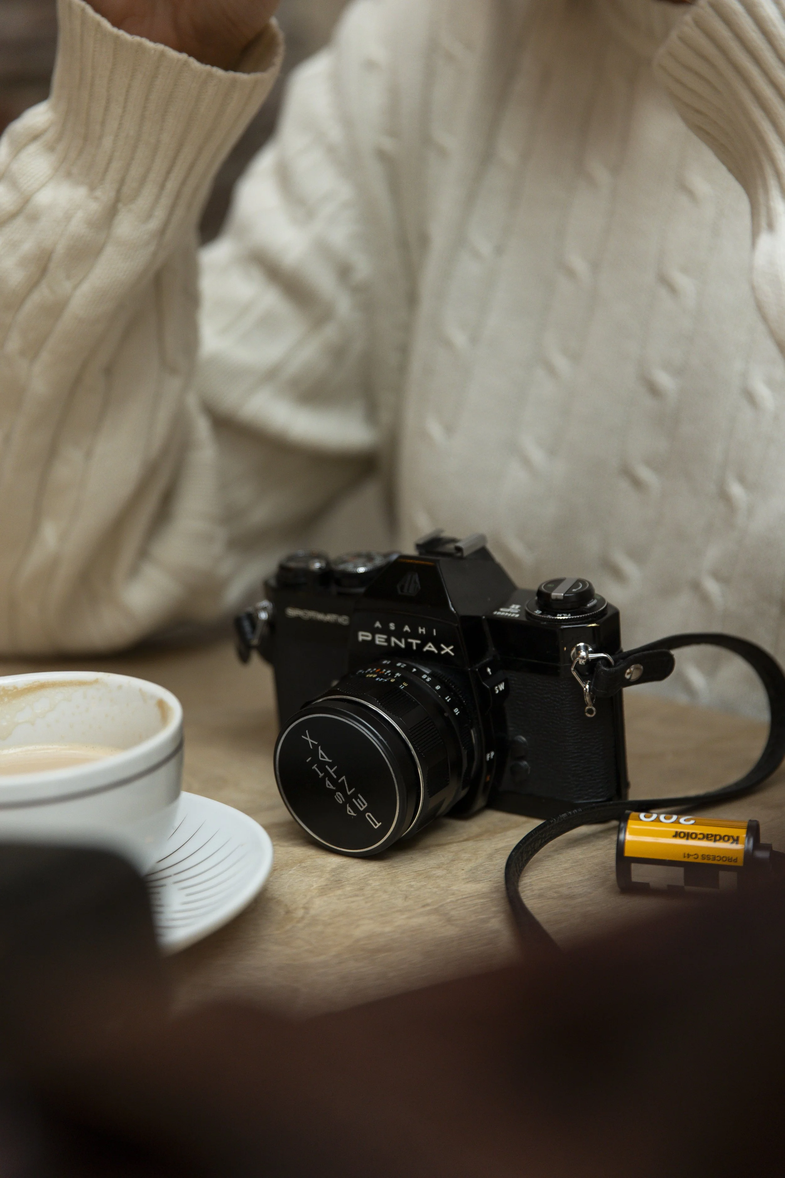 Close-up of a vintage black Asahi Pentax camera, a partially visible cup of coffee, and a yellow Kodak AA battery on a wooden table, with a soft beige sweater in the background.