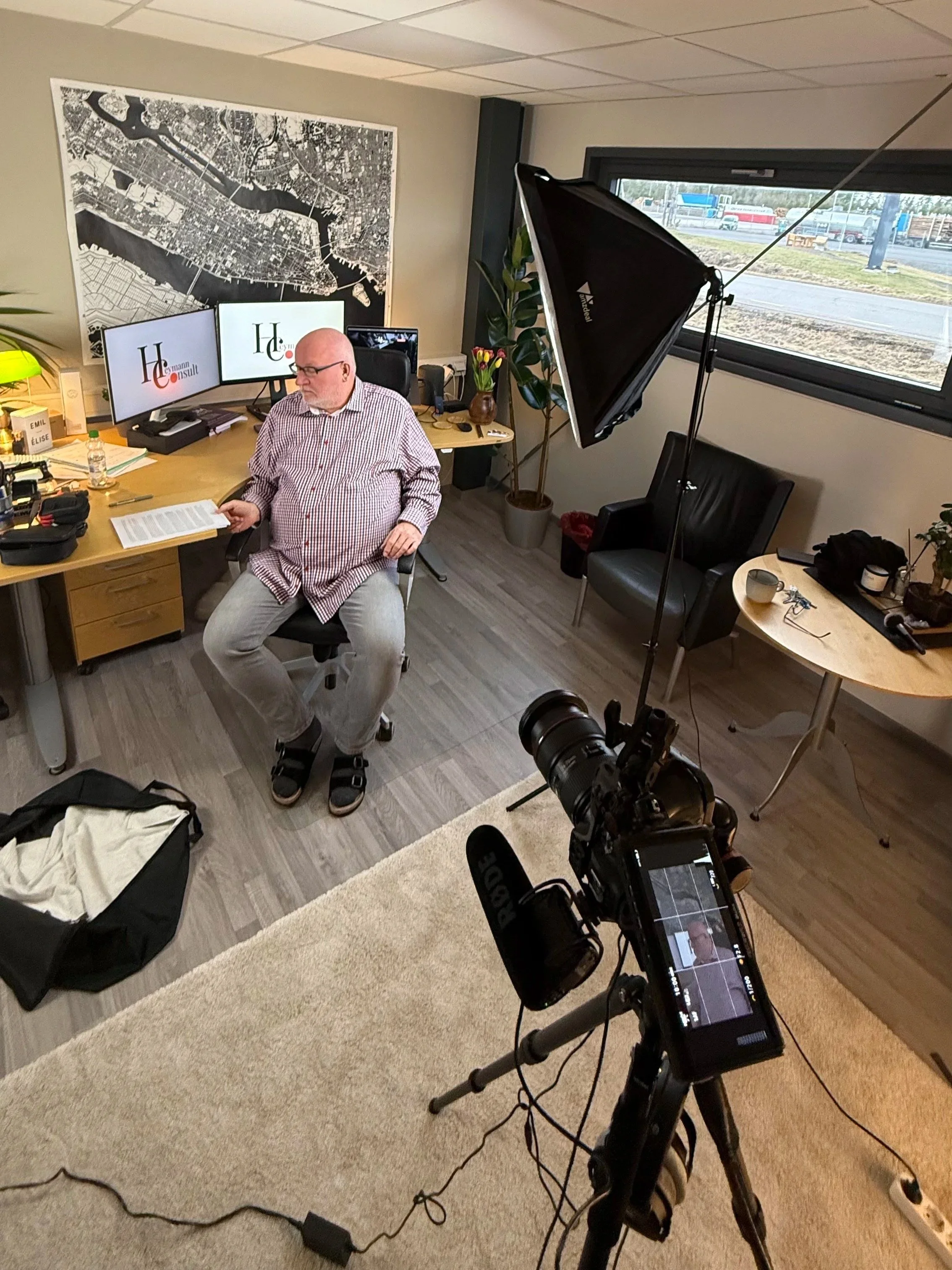 A man sitting in an office near a desk with two computer monitors, a potted plant, and various office supplies. Photography equipment, including a large softbox and a camera on a tripod, is set up nearby. A window shows a highway with vehicles outside.