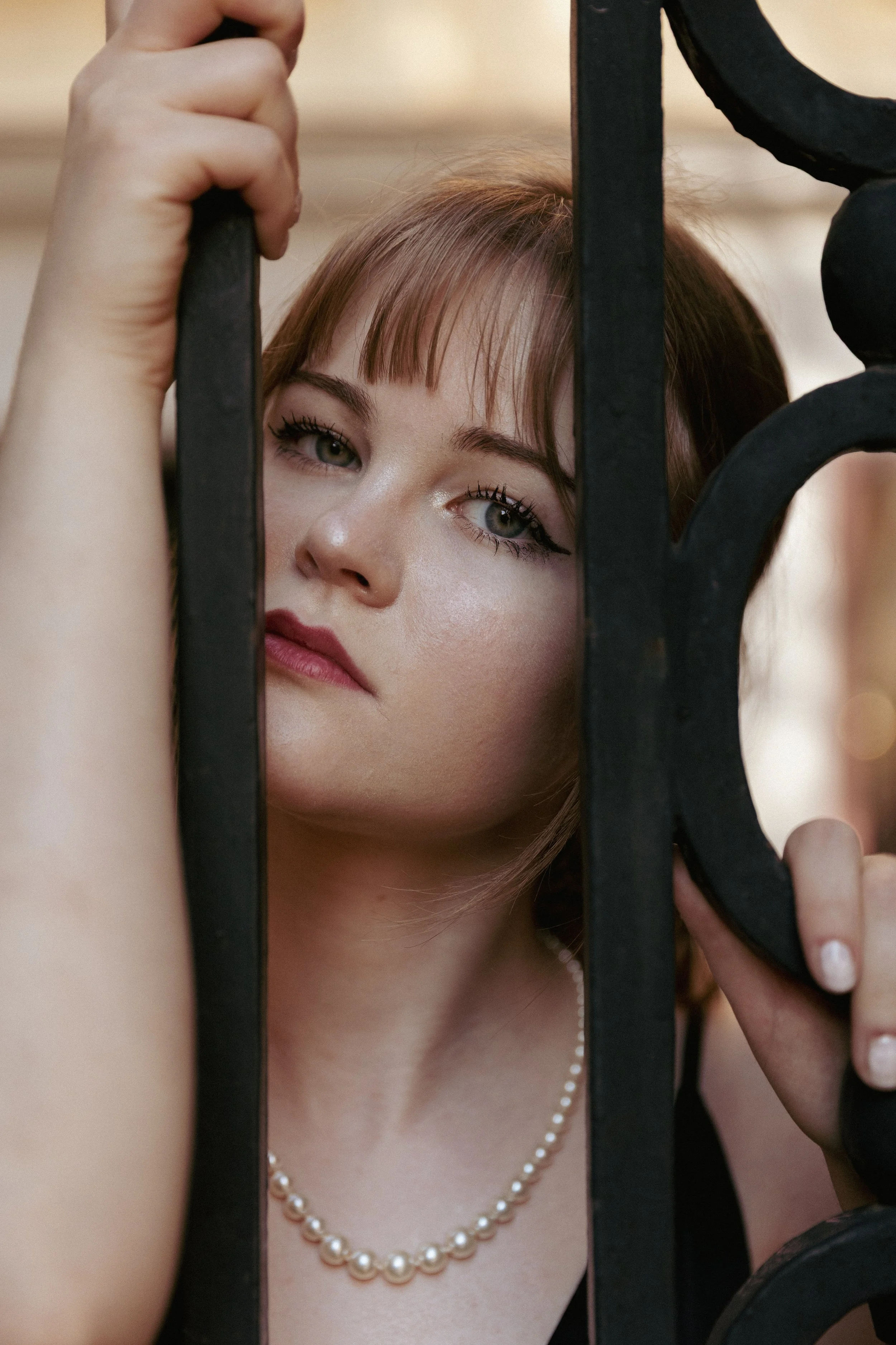 A woman with light skin and shoulder-length brown hair, wearing a string of pearl necklace, looking through black iron bars.