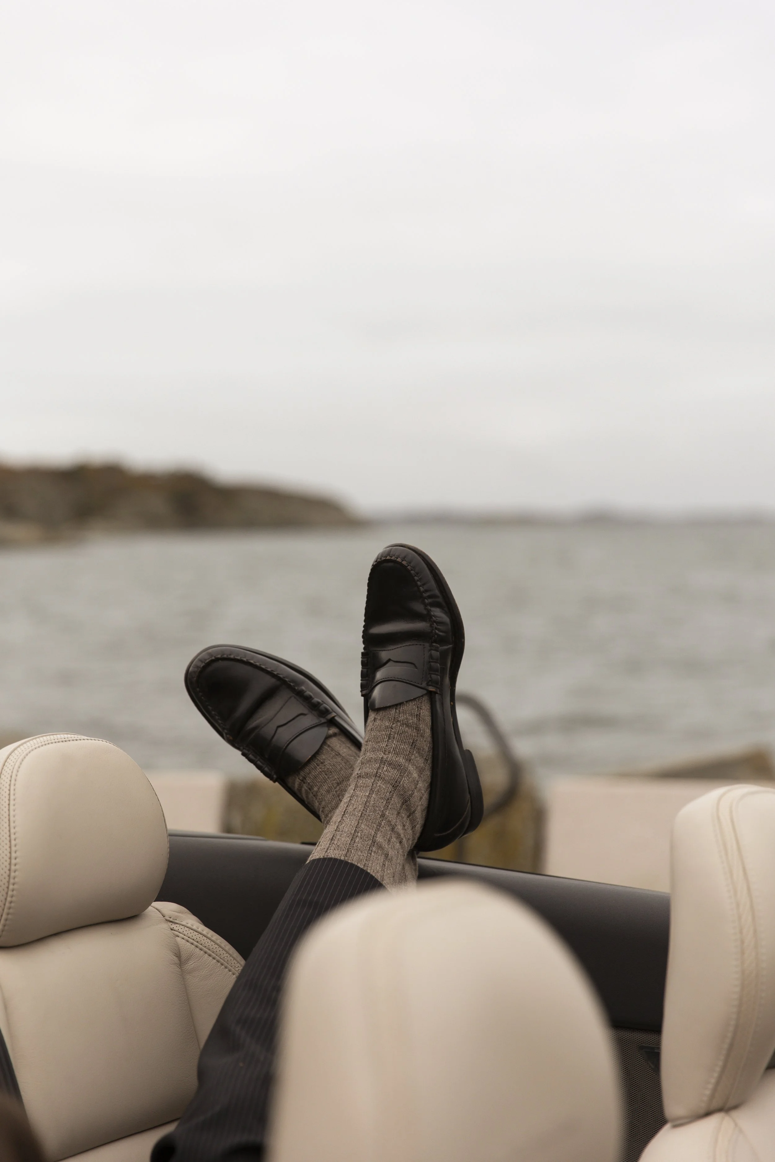 Person relaxing in a luxury convertible car with cream-colored leather seats, wearing black shoes and gray pants, with legs resting on the car's backrest, overlooking a scenic body of water and distant landmass under an overcast sky.