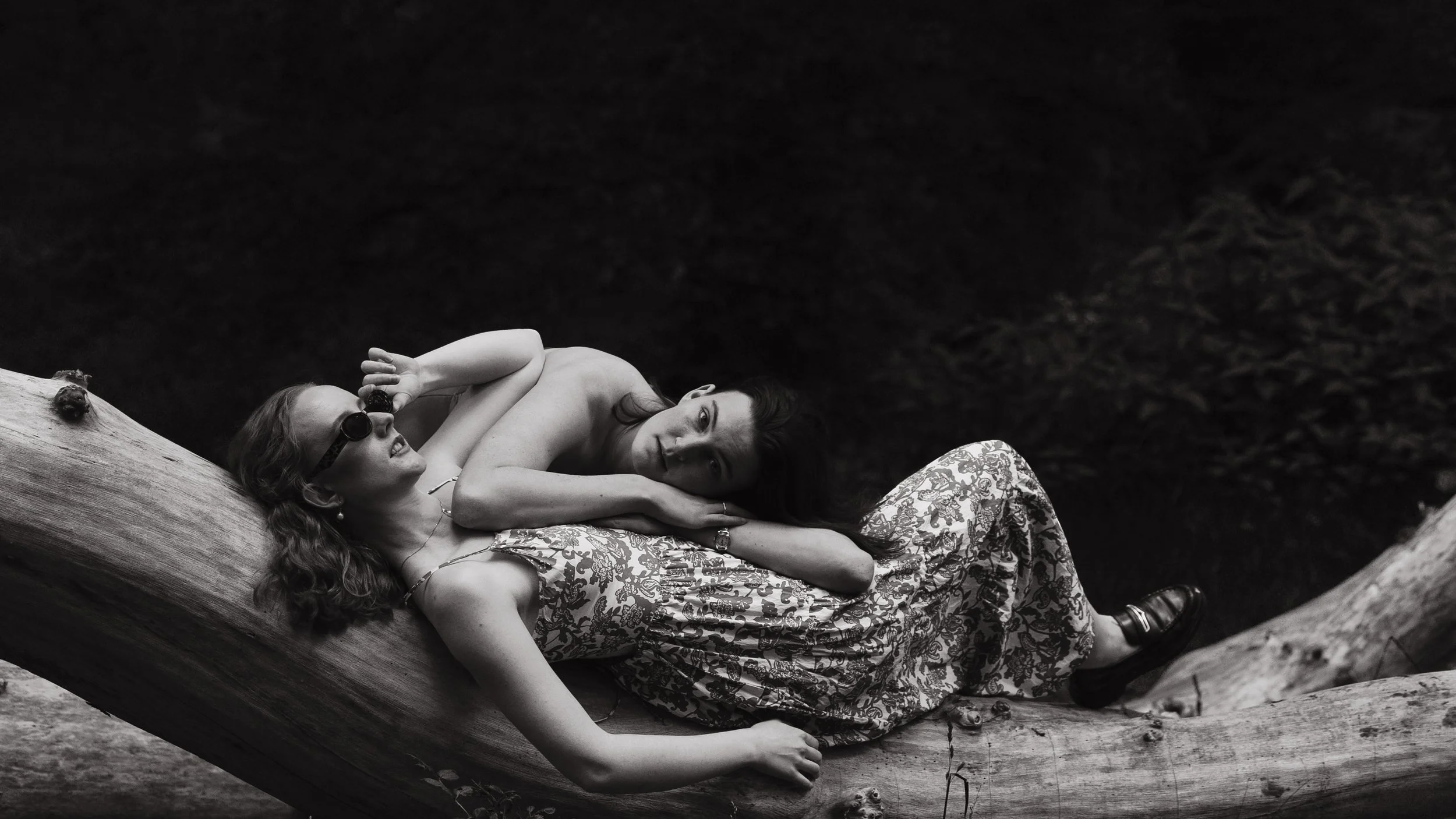 Two women relaxing on a large fallen tree trunk by a body of water, with one woman wearing sunglasses and the other lying on her side, gazing at the camera.