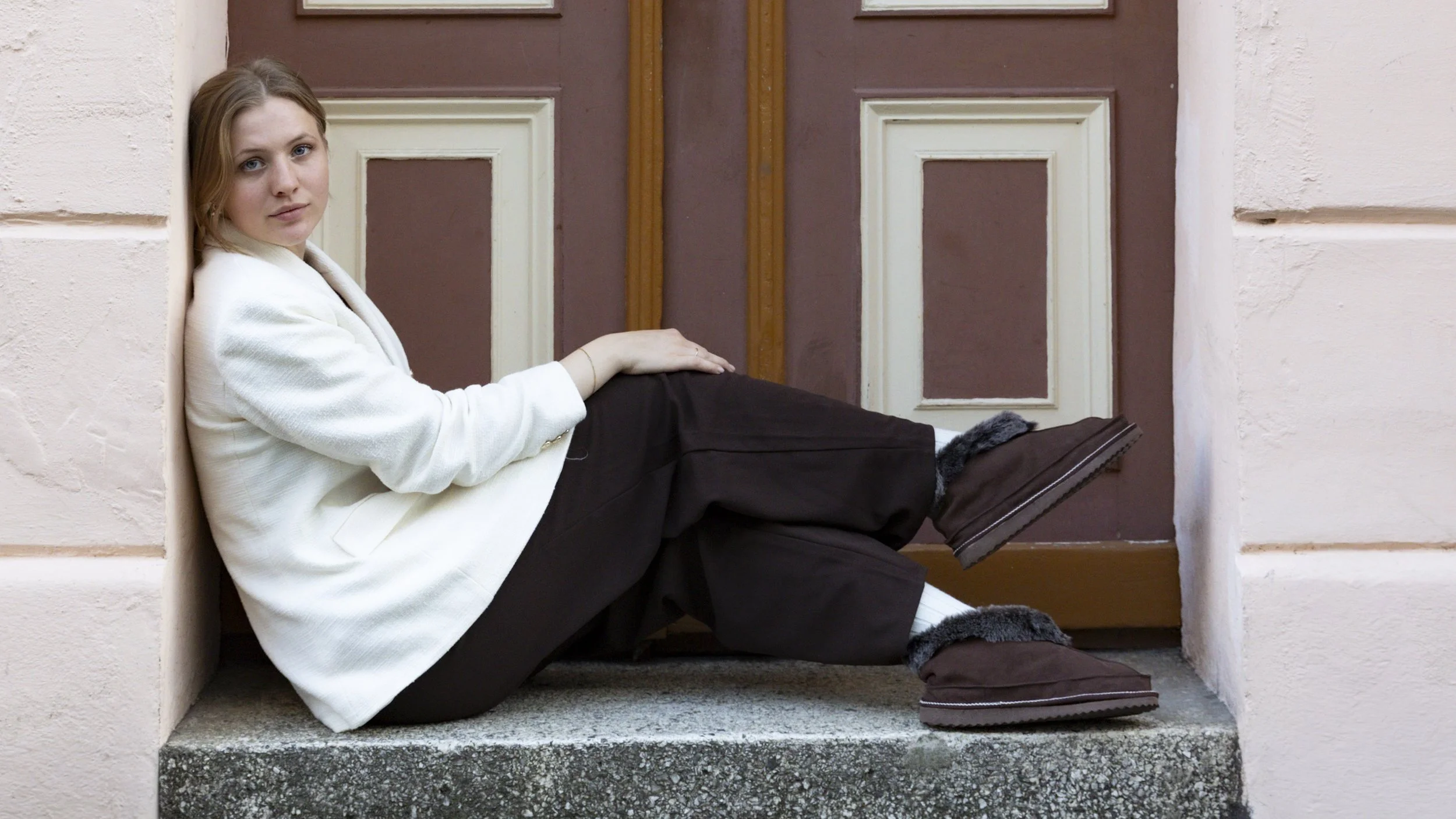 A young woman with light brown hair and blue eyes sitting on a windowsill, leaning against a wall, with her legs stretched out in front of her. She is wearing a white jacket, dark pants, and brown fur-lined boots, and looks directly at the camera.