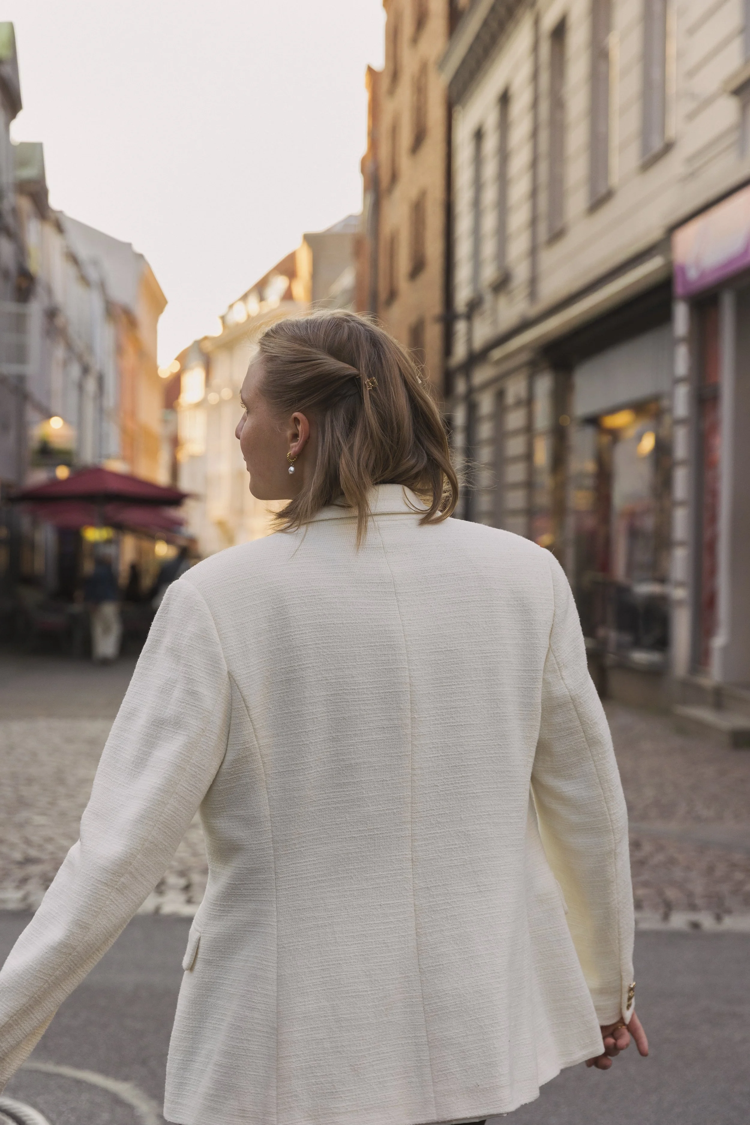 A woman wearing a white coat and pearl earrings walking down a cobblestone street with buildings on either side.