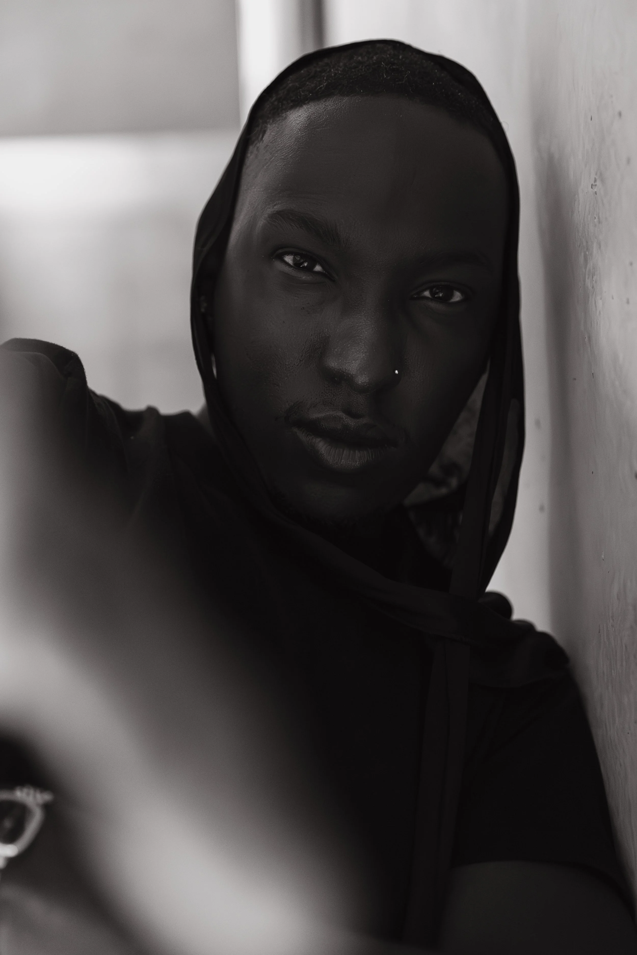 Close-up black and white photo of a person's face with a headscarf, leaning against a wall, looking directly at the camera.