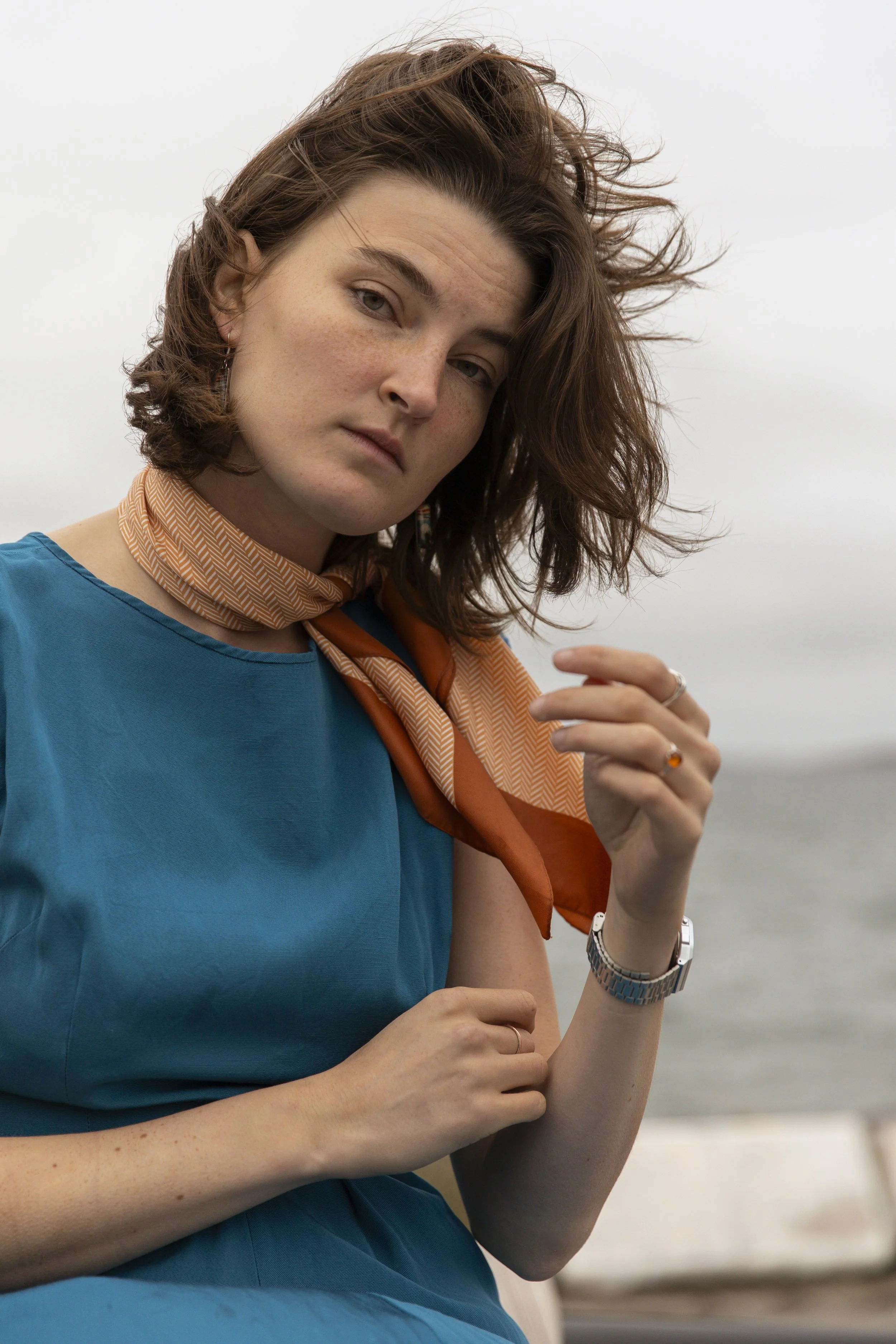 A woman with short, curly brown hair and light skin is looking directly at the camera with a neutral expression. She is wearing a blue top and an orange patterned scarf around her neck. She has a silver watch on her left wrist and a ring on her right ring finger. The background is a cloudy sky.