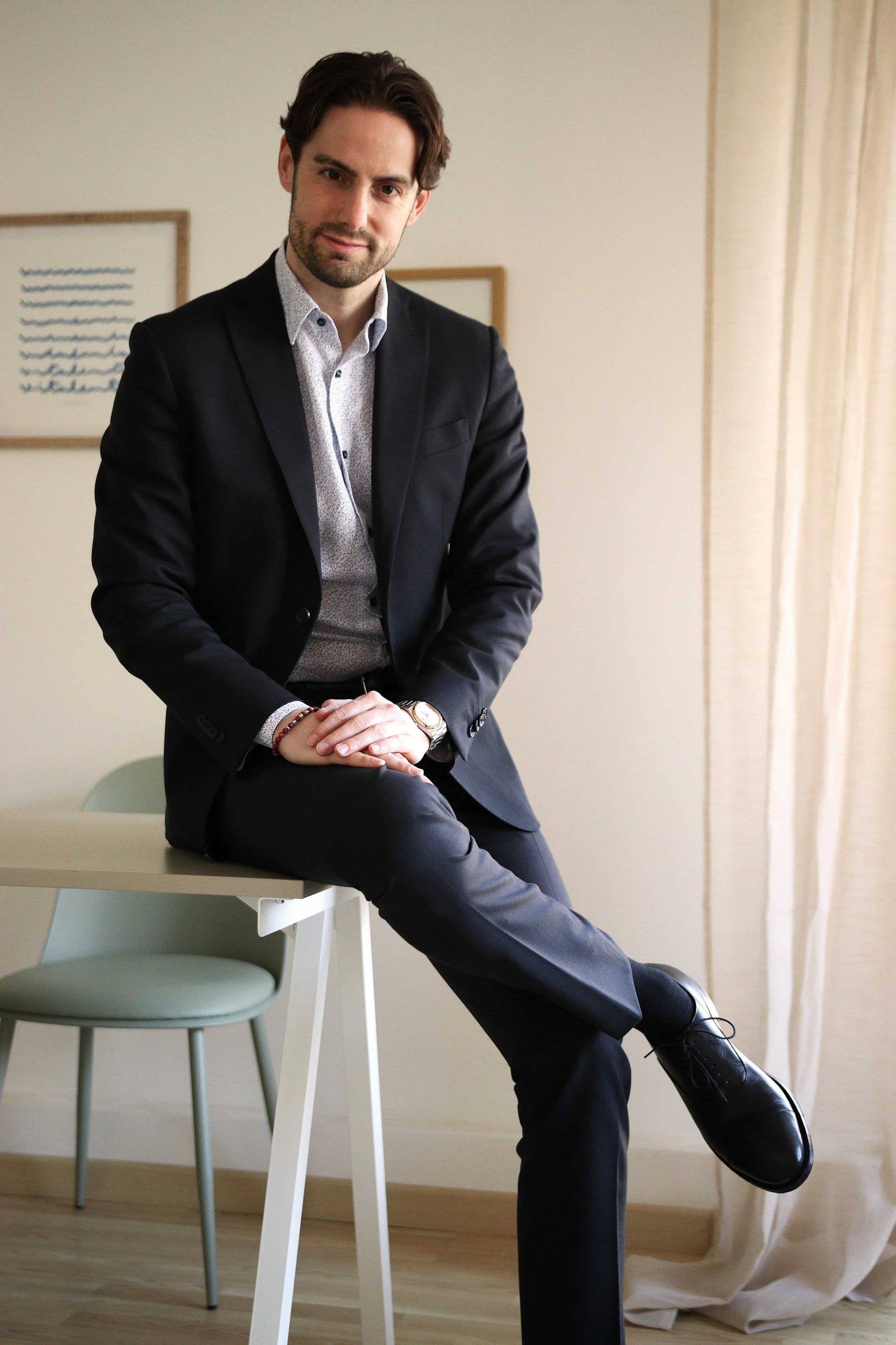 Psicologo seduto su un tavolo con gambe incrociate, vestito elegante, in una stanza luminosa con tende chiare e quadri alle pareti.