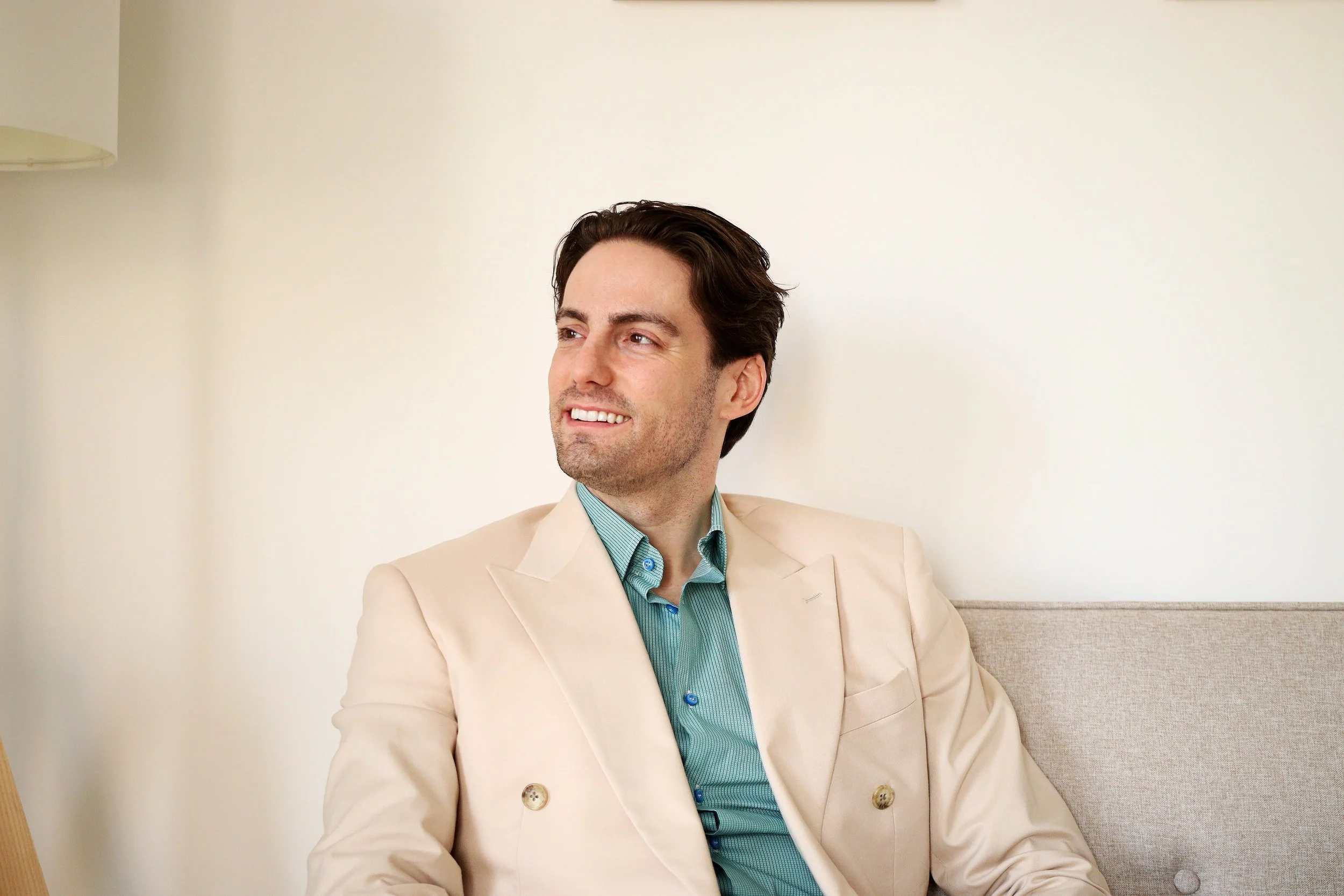 Psicologo esperto in autostima e gestione dello stress, con capelli scuri e sorriso, indossa un completo beige e una camicia verde, seduto su un divano contro una parete bianca.