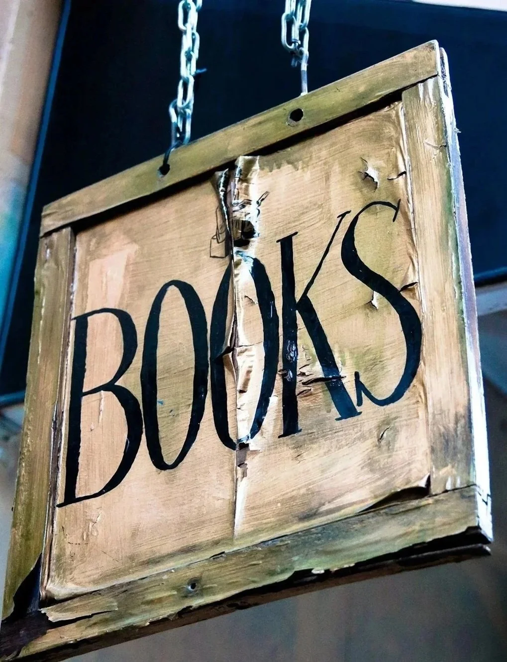 Wooden sign with the word 'BOOKS' written on it, hanging from chains, with visible damage and tears in the wood.