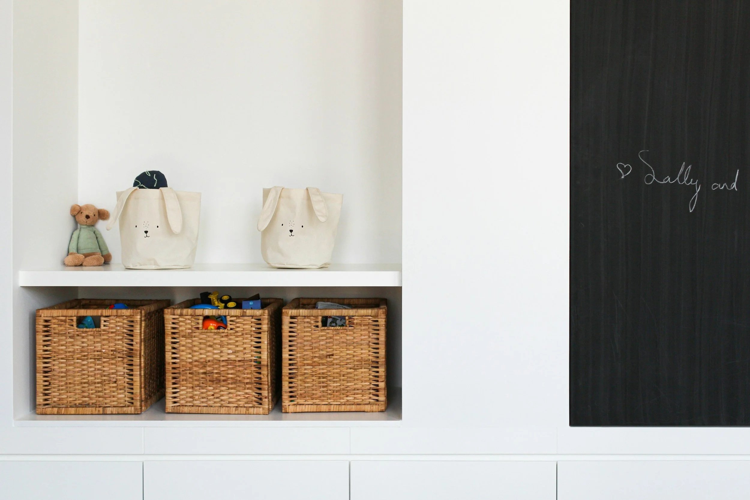 White shelves with three woven baskets containing toys, a stuffed bear, and two tote bags with animal faces, a blackboard with the words "Sally and" written on it.