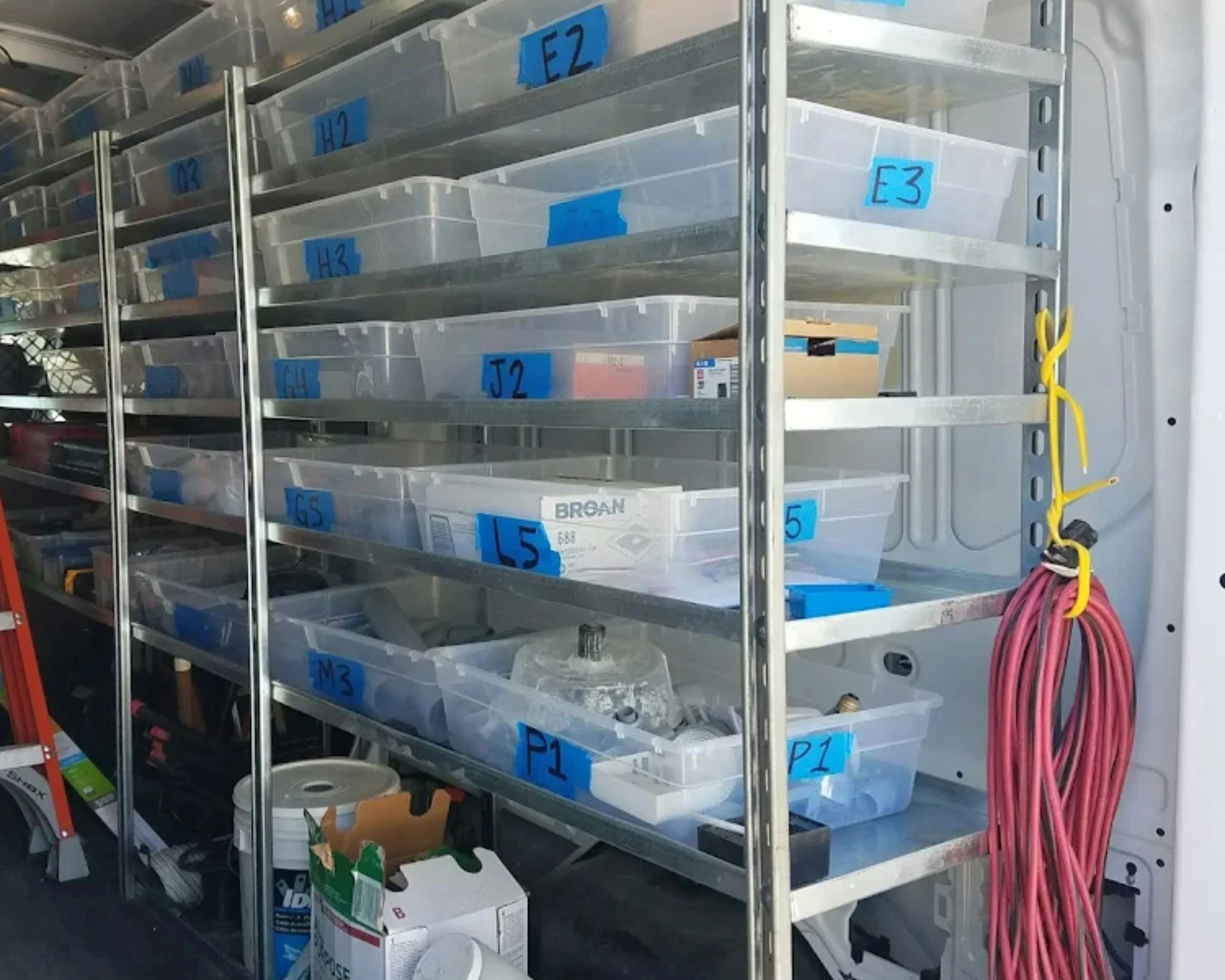 A metal shelving unit with multiple clear plastic bins labeled with blue tape, containing various small parts and supplies. Power cords are hanging on the right side of the shelf.