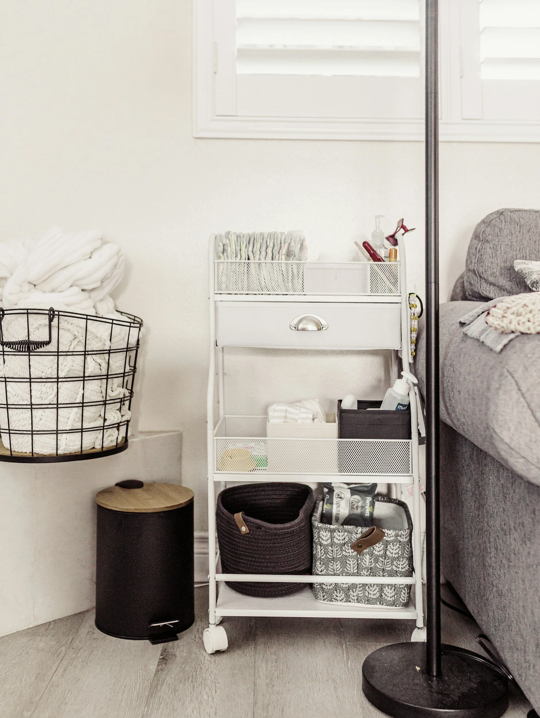 A white rolling cart with multiple shelves holding various small items, situated next to a couch with a pillow and blanket, a black wastebasket, a black floor lamp, and a basket filled with blankets on the floor, in a room with white walls, a window, and light wooden flooring.