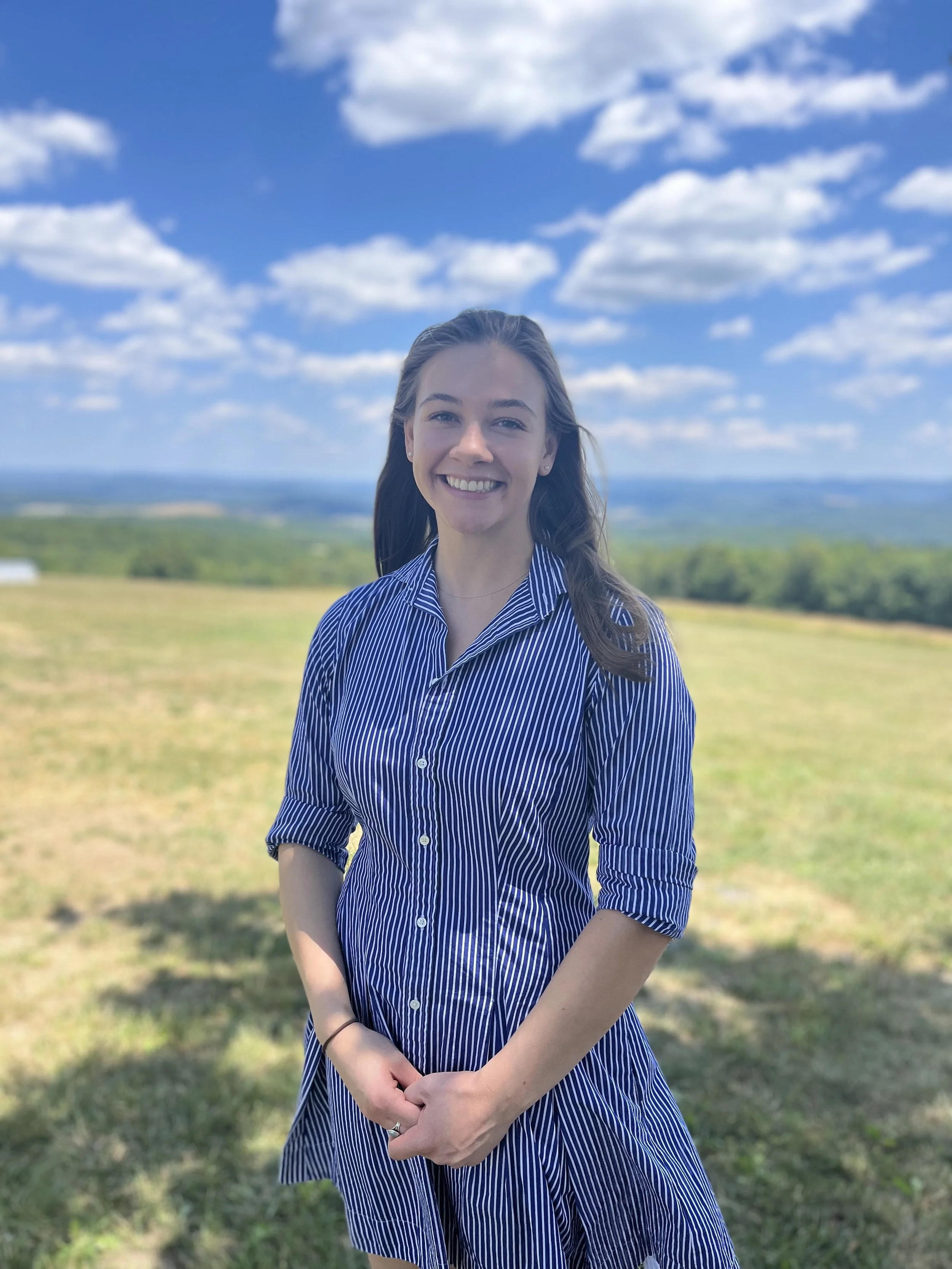 A young woman with long brown hair smiling outdoors, standing in a grassy field with trees and a blue sky with scattered clouds in the background.