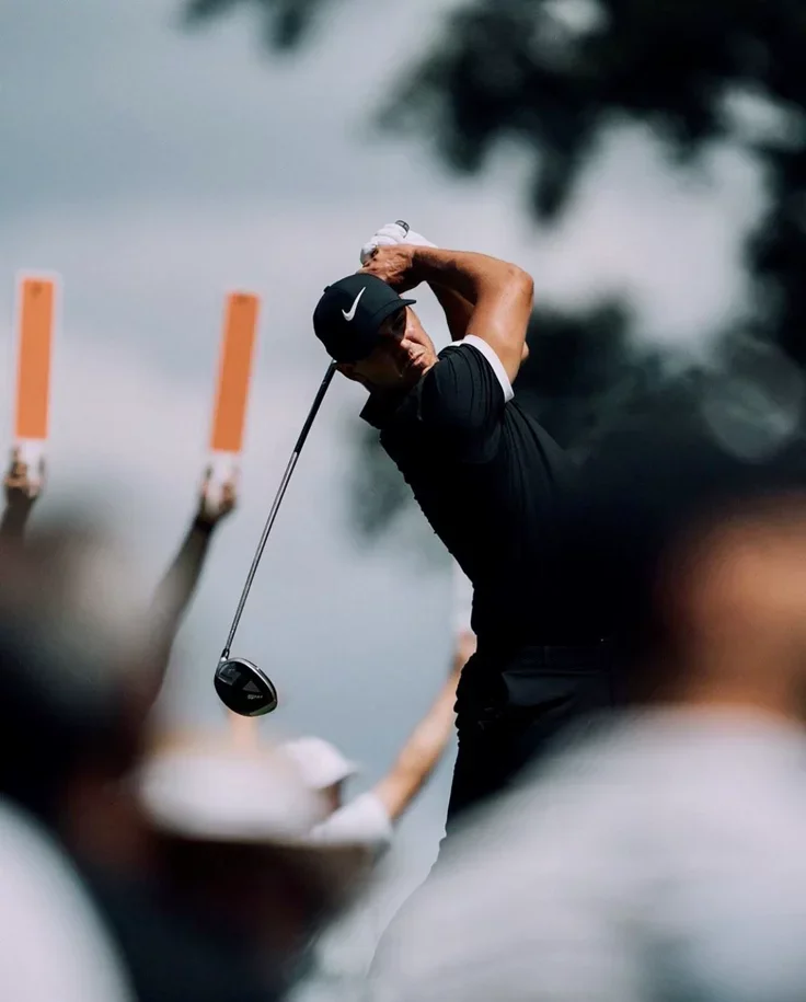 A golfer in black attire and a Nike cap swings a golf club on a golf course.