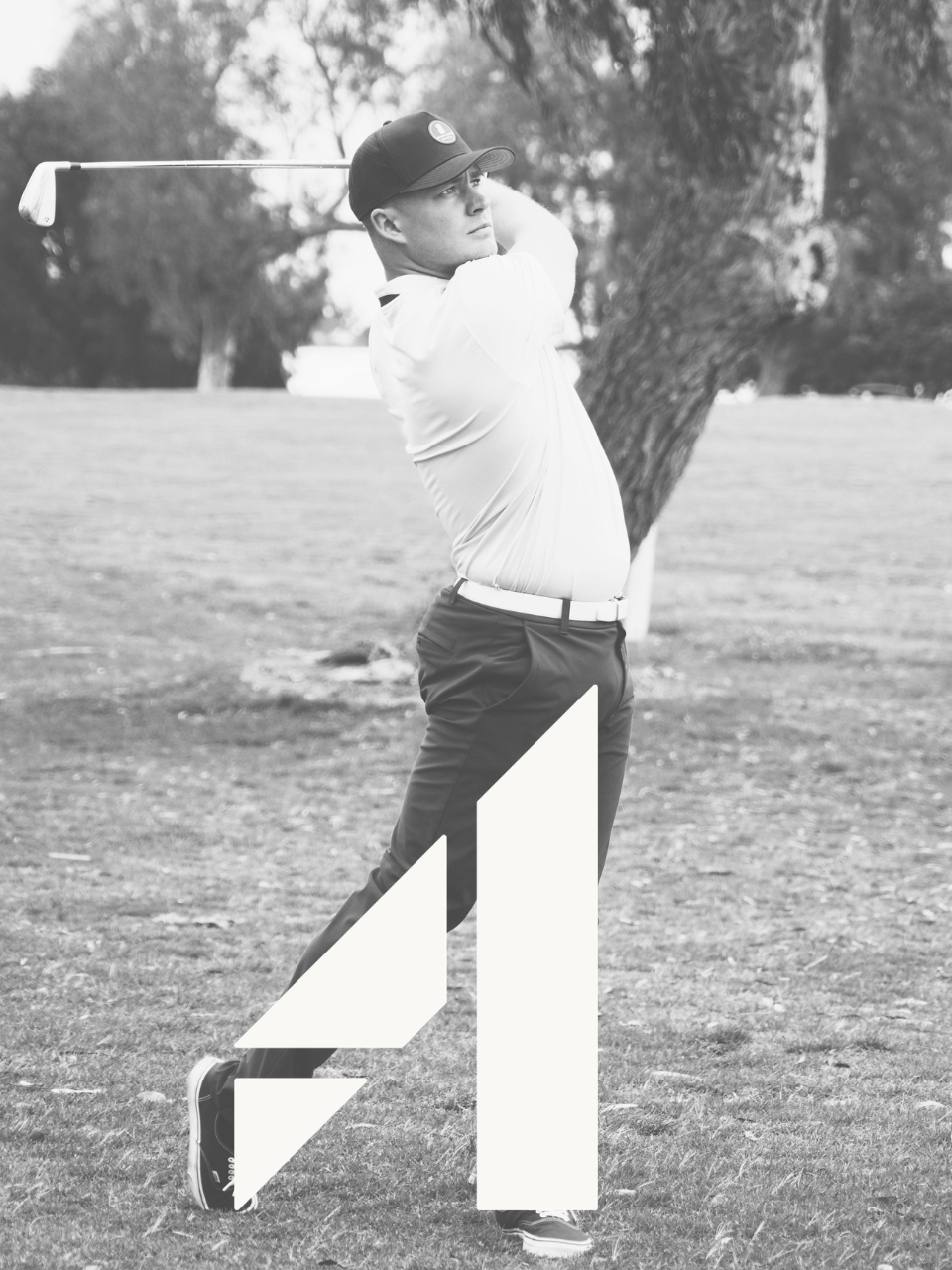 A man playing golf, swinging a club on a golf course, wearing a cap, white shirt, and dark pants.