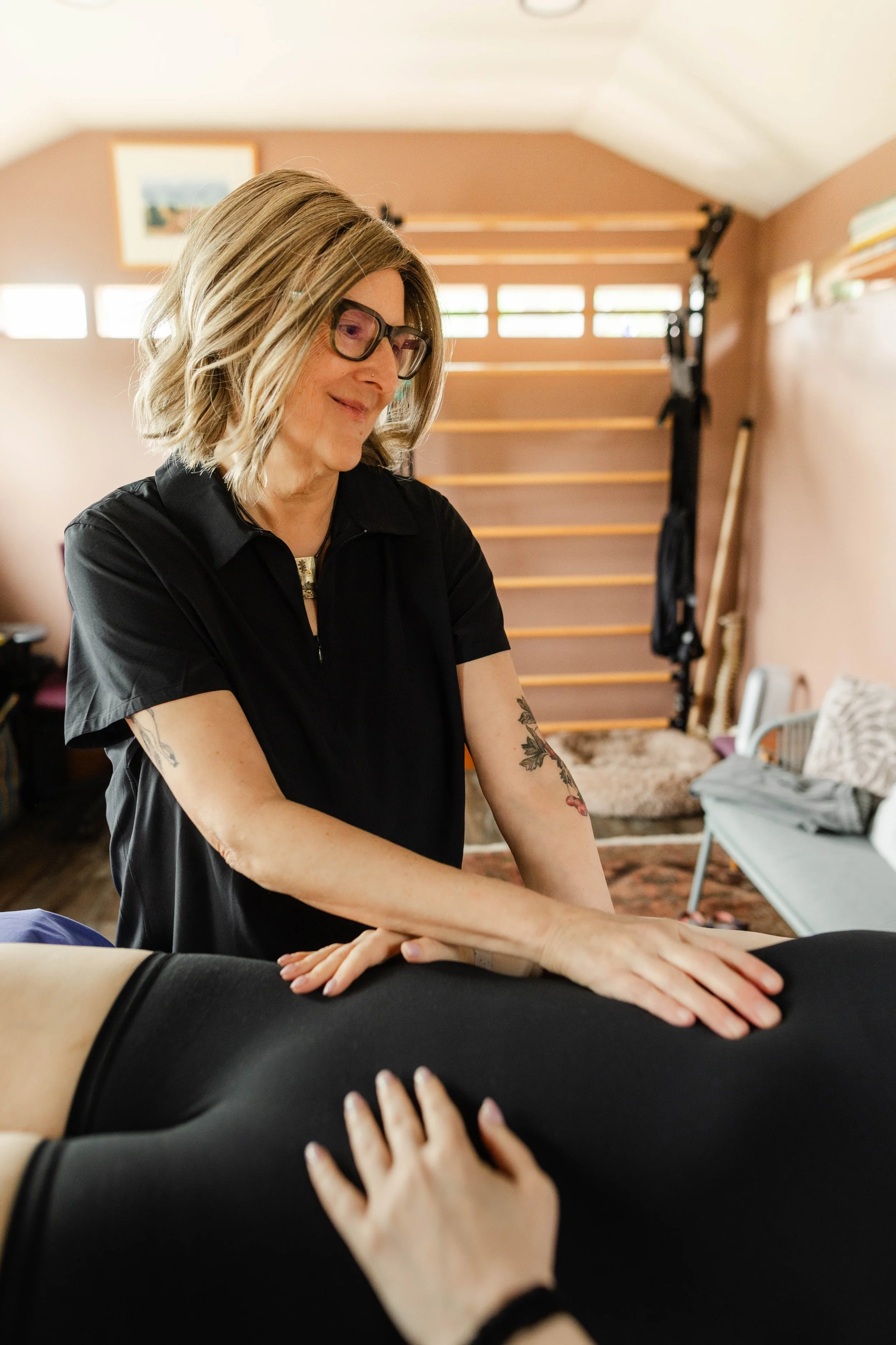 A woman with shoulder-length blonde hair, glasses, and tattoos on her arms is providing bodywork to someone lying face down on a massage table.