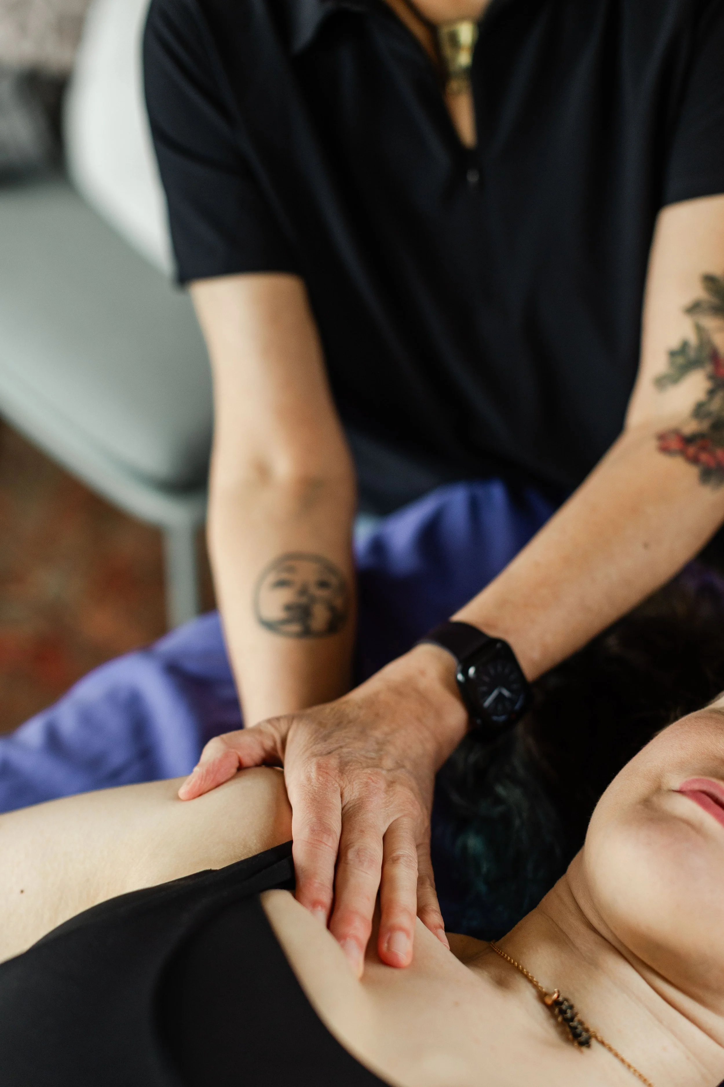 A woman providing a Rolfing session.The woman is wearing a black top, a necklace, and has a tattoo on her arm.