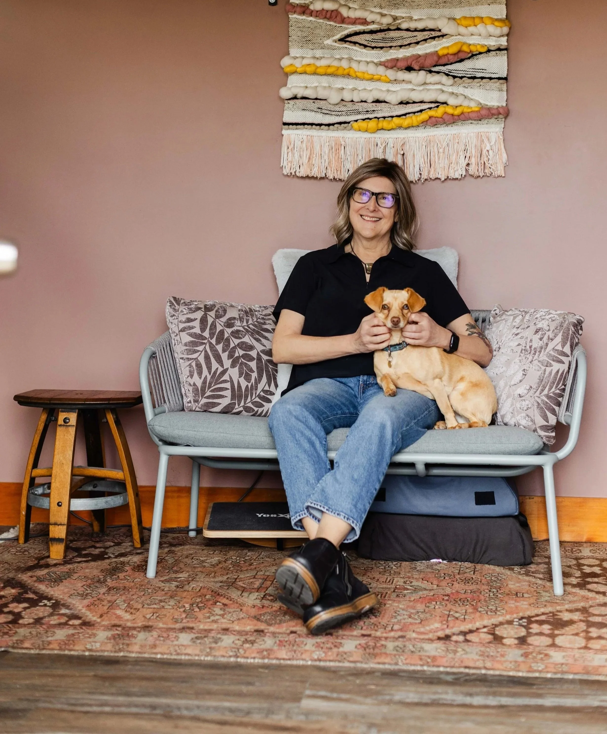 A Rolfing practitioner with glasses and shoulder-length hair sitting on a gray sofa with patterned pillows, holding a small tan dog, in a room with a pink wall and a textile wall hanging. There is a small wooden side table to her left.